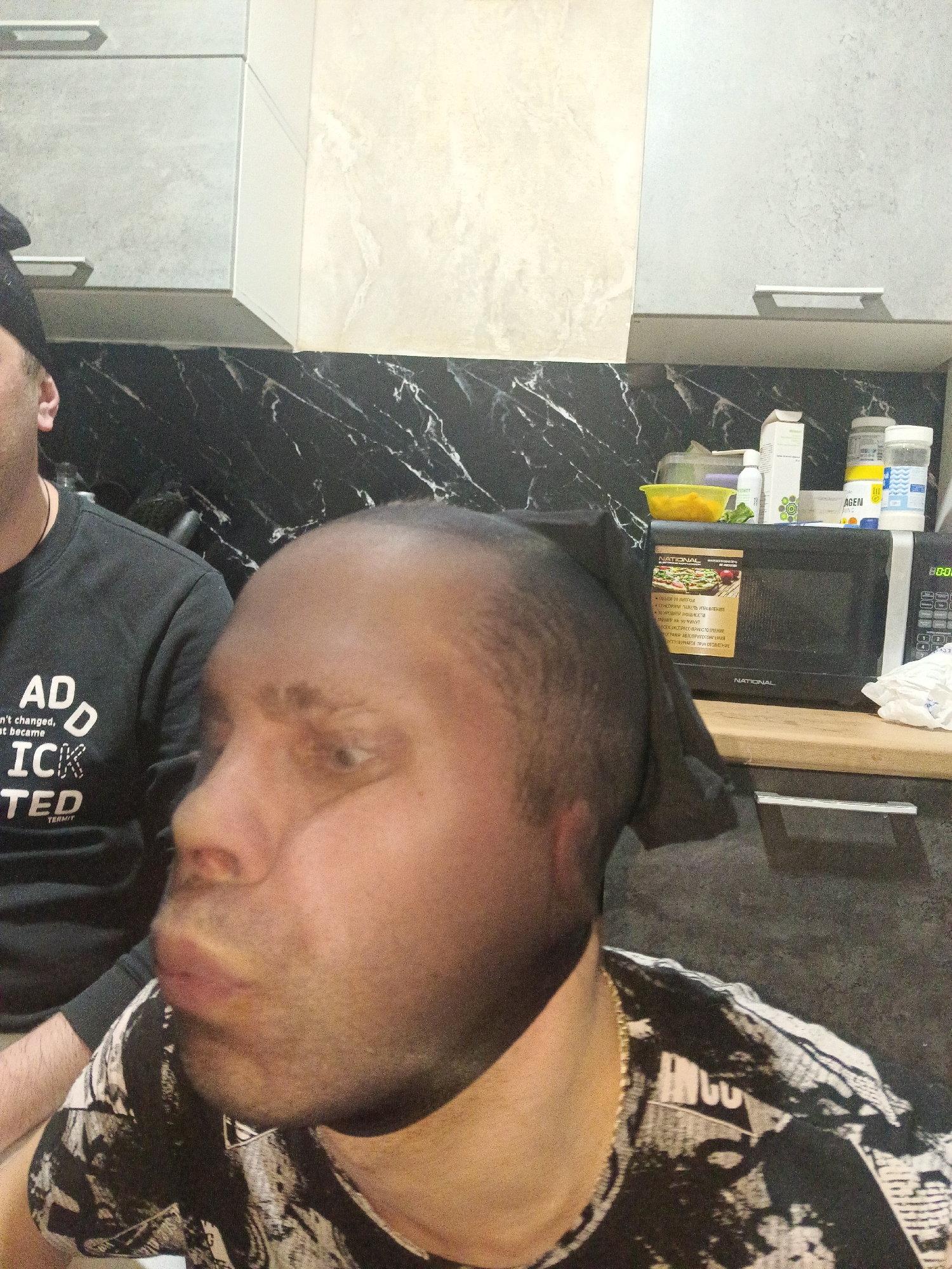 Two men are in a kitchen in what may be a private residence in Chaikovsky, Russia. One man is wearing a black sweatshirt with the words "ADD...ICK TED" on it. The second man is wearing a t-shirt that is black and white and a thin gold chain. In the background, there are black marble countertops and grey and white cabinets. A microwave, brand "NATIONAL" is on the counter, with a yellow container, bottles, and collagen supplements on top of it. A roll of paper towels is next to the microwave.