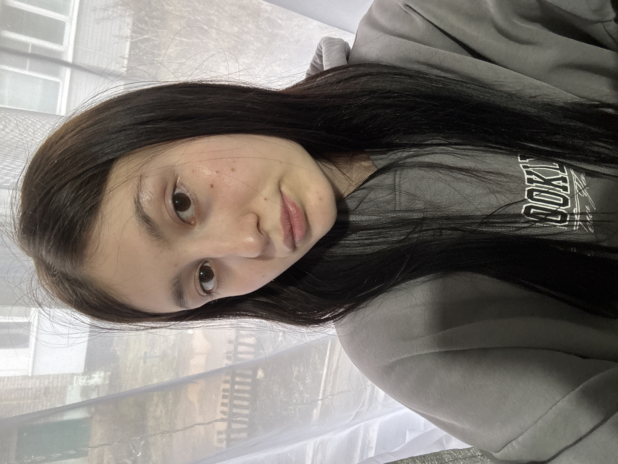 A portrait-oriented photograph, taken sideways, features a young female subject indoors. She has long, dark, straight hair flowing around her shoulders and chest. Her face has a neutral to slightly pensive expression, with dark eyes looking towards the camera and her lips slightly parted. Her skin shows a few natural marks or freckles.
She is wearing a gray hooded sweatshirt. Visible white text on the front of her sweatshirt includes "OKL" and "LYN", suggesting the word "BROOKLYN".
The background consists of a window with sheer white curtains or blinds, allowing diffused daylight to enter the room. Through the translucent curtains, blurry outlines of distant buildings or structures are discernible, indicating an urban or suburban setting outside. The natural lighting suggests the photo was taken during the daytime. This scene is located in Фоки, Russia.