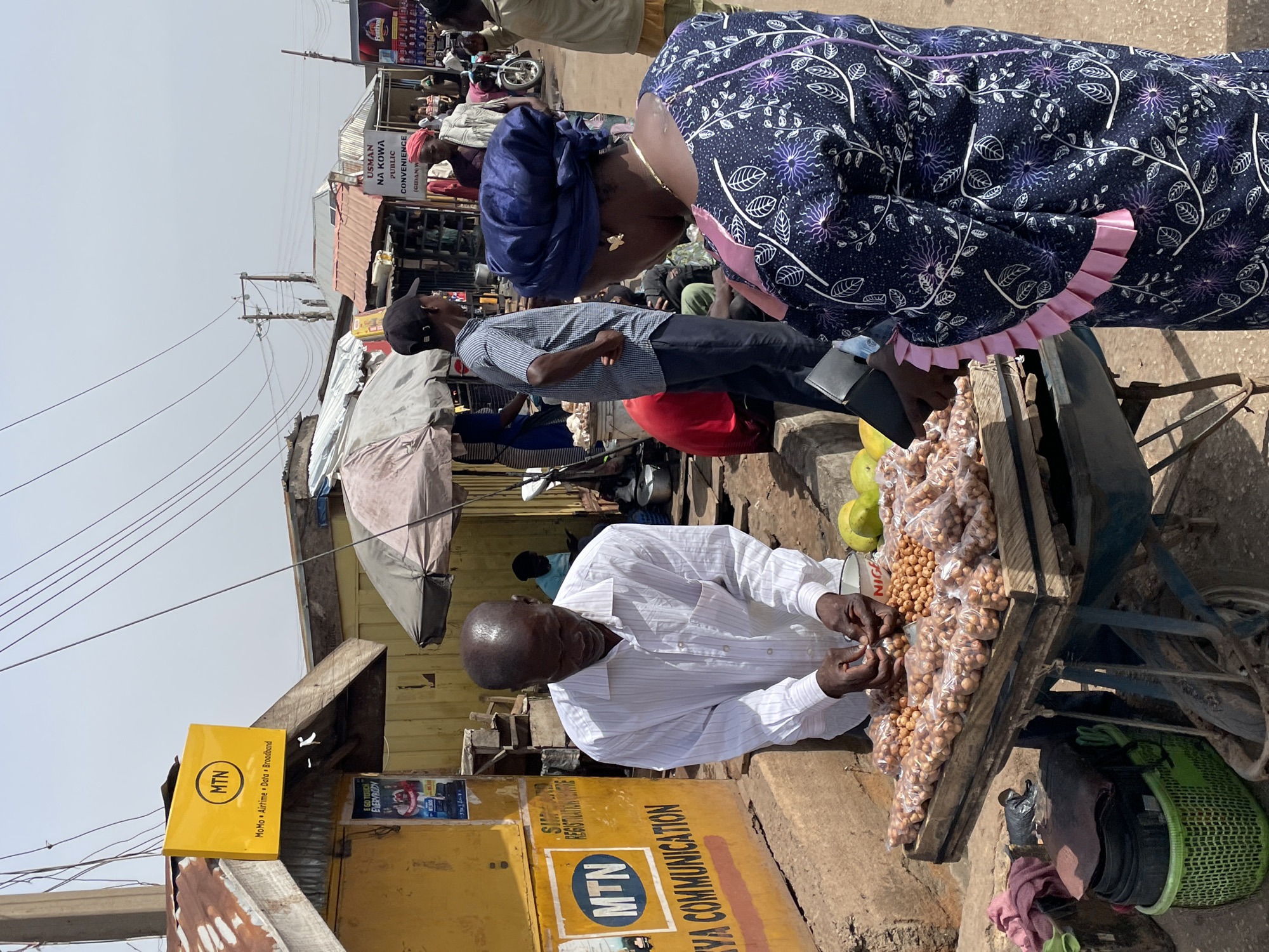 The image captures a street scene in Jos, Nigeria, bustling with activity. A bald, elderly man in a white, long-sleeved shirt is selling peanuts from a wooden cart laden with plastic bags full of the snack. Two customers, a woman in a navy blue patterned dress with pink ruffles and a woman with a dark blue head covering, are interacting with him. A man in a blue shirt and dark pants stands behind the women.
In the background, a yellow MTN (Mobile Telecommunications Network) sign is visible on a building. Other signs and posters are present, including one that reads "USMAN NA KOWA PUBLIC CONVENIENCE." People can be seen milling about in the background. Power lines crisscross the sky. The scene appears to be a typical marketplace or roadside vending area.