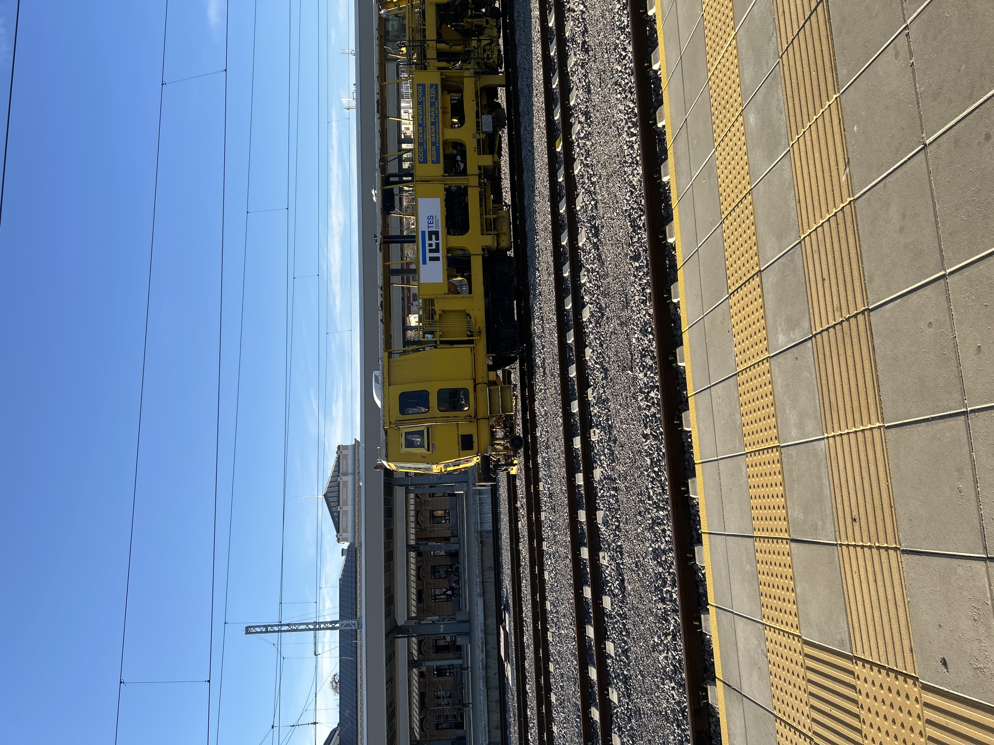 The image shows a section of a railway station in Plovdiv, Bulgaria. A yellow railway maintenance vehicle is parked on the tracks next to a platform. The platform is made of concrete slabs with tactile paving featuring raised bars and dots. Overhead power lines run parallel to the tracks. Behind the yellow vehicle is a building that is part of the train station infrastructure. The sky is clear and blue.