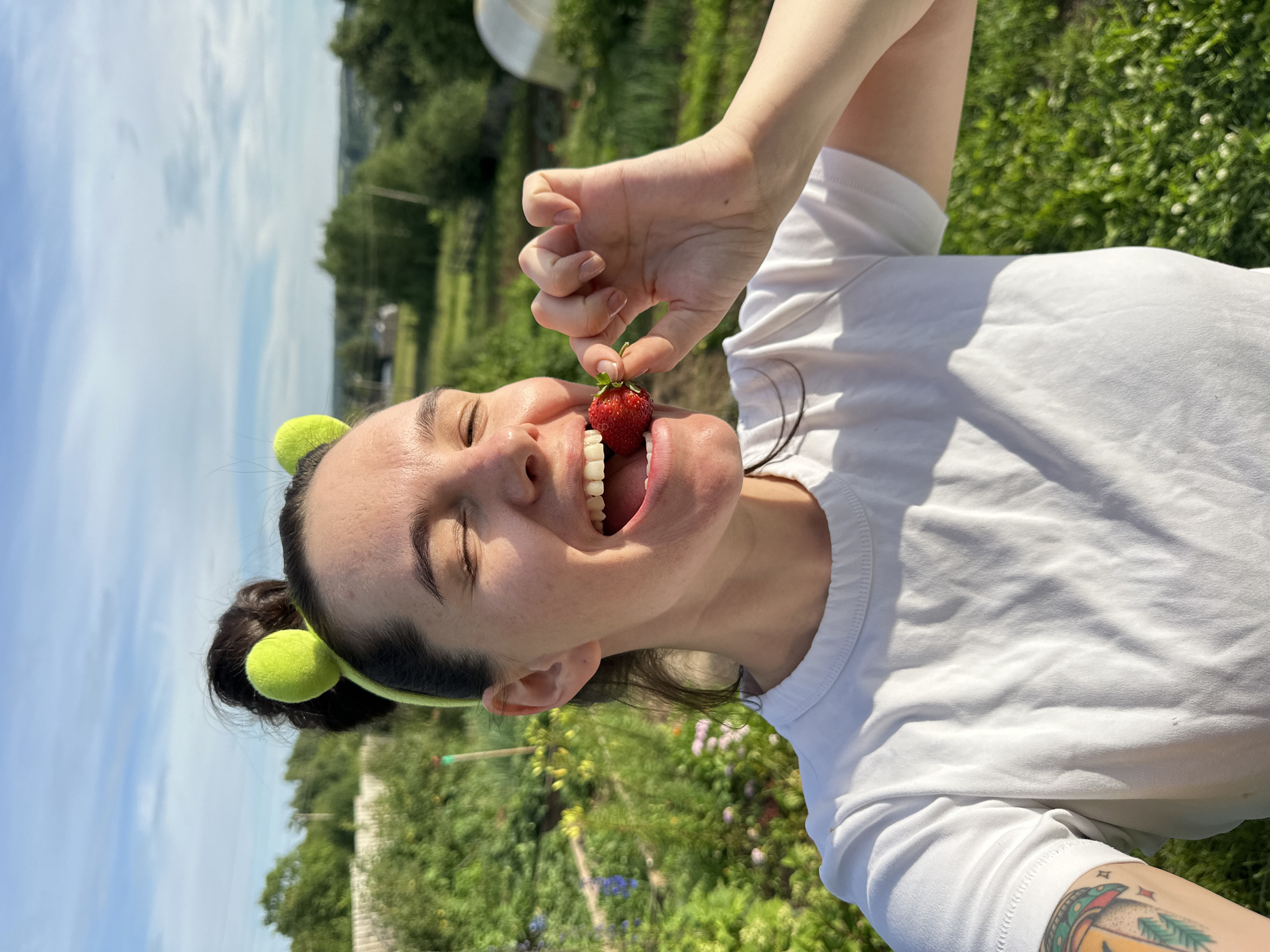 In a sunny outdoor scene, a woman with her eyes closed and mouth open is about to eat a strawberry. She is holding the strawberry with her right hand, bringing it to her mouth. She is wearing a white t-shirt and a green headband with two fuzzy balls on top. The background is a lush green field with trees and a bright blue sky with some clouds, suggesting it is daytime with good weather. The woman appears joyful and is likely in a rural or garden setting in Альняш, Russia. A tattoo is visible on her arm.