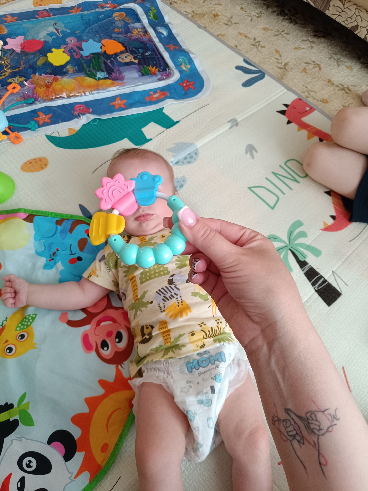 A baby lies on a play mat with cartoon animals and dinosaurs. The baby wears a patterned shirt and a diaper labeled "Momi." A hand holds a colorful teething toy in front of the baby's face. Another person's legs are visible in the upper right corner. The photograph's location is Tobolsk, Russia. A tattoo is visible on the arm holding the toy.