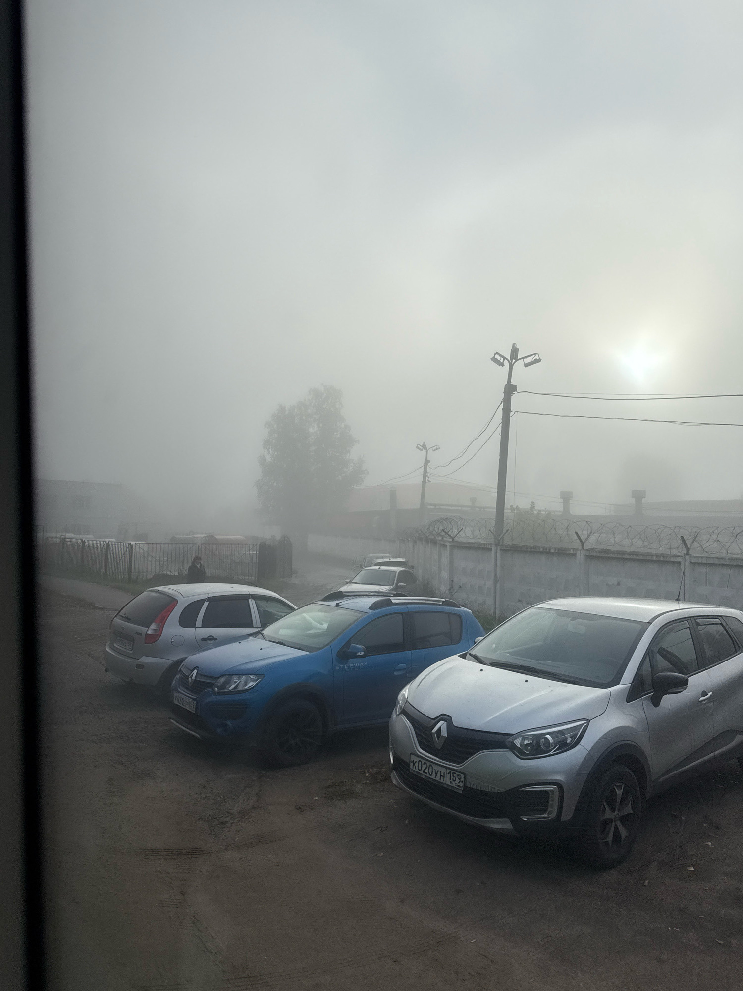 An outdoor, low-angle, slightly blurred photograph captures a foggy street scene in Chaikovsky, Russia. The foreground features a dirt or gravel parking area with three cars: a silver Renault Captur in the front right, a blue Renault Sandero Stepway in the middle, and a silver Lada Kalina in the middle left. The Renault Captur has its license plate visible, reading "K020YH 159". Behind the cars, a concrete fence topped with barbed wire runs horizontally across the frame, separating the parking area from a factory or industrial complex. A person wearing dark clothing is partially visible standing behind the Lada Kalina, near the fence. In the background, trees are obscured by dense fog, and a utility pole with lights is visible to the right, silhouetted against a hazy sun. The overall atmosphere is overcast and damp.