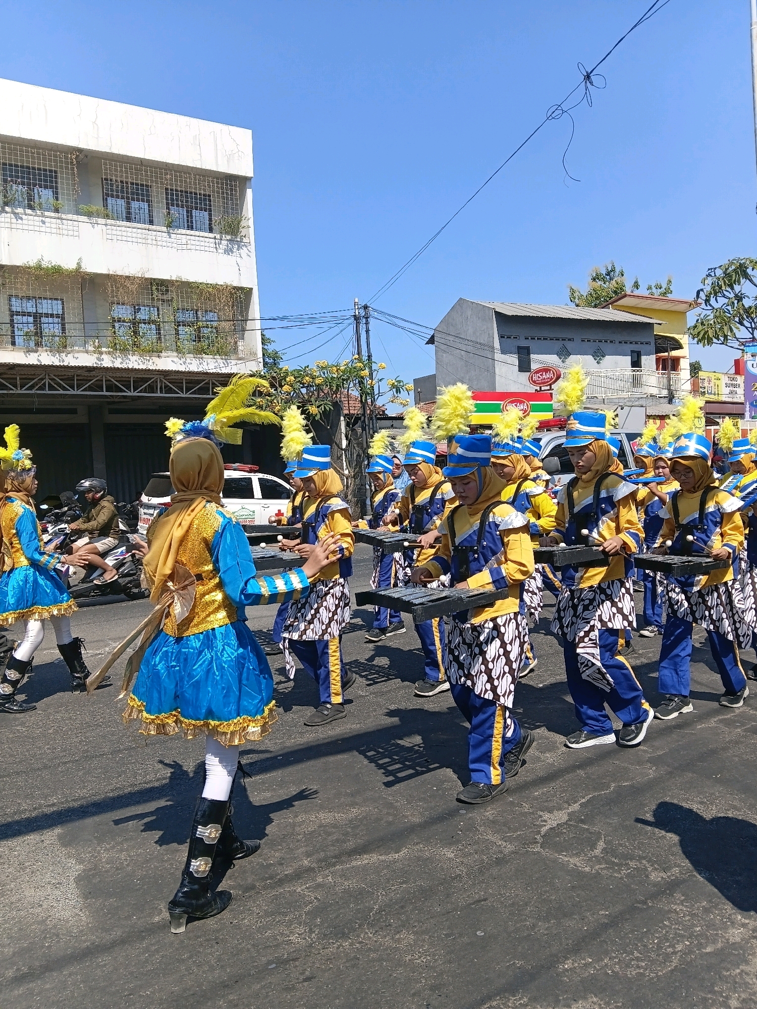 In Wadungasri, Indonesia, a parade is underway on a sunny day. A group of individuals, appearing to be predominantly young women, march in unison, dressed in elaborate blue and gold marching band uniforms adorned with yellow feathers. They are playing metallophones. The parade route is paved, with the road showing signs of wear. On the left side of the street, a white building with windows protected by metal grates stands, with some vegetation growing on it. To the right, houses and businesses are visible, including a red and white sign for "HISANA" and another for "TOKO SUMBER JAYA". Several motorcycles are parked or being ridden on the side of the road, and a white vehicle, possibly an ambulance, with red and blue lights is also present. The sky is clear and blue, indicating fair weather. The people are focused on their performance, moving forward in formation.