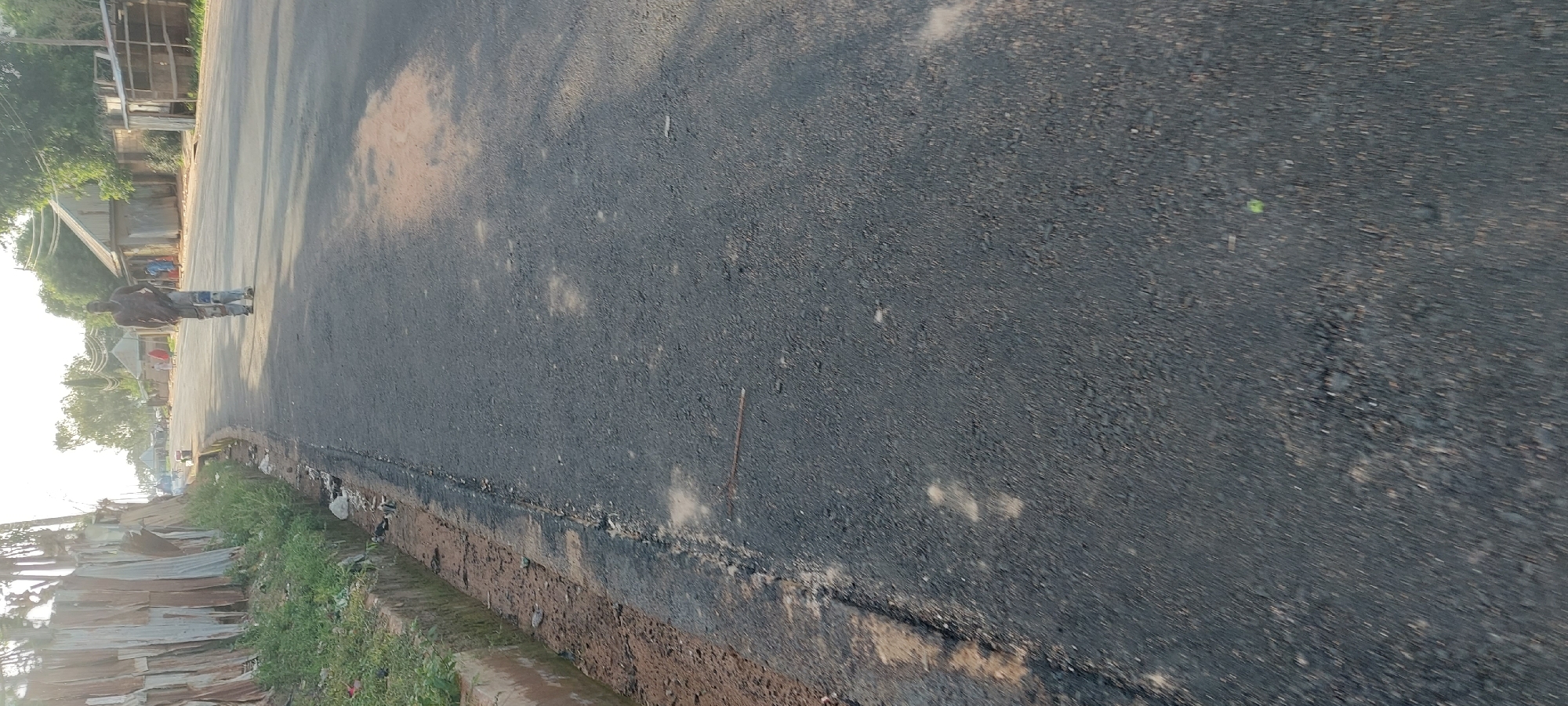 The image shows a man walking on a paved road in Mayo Dasa, Nigeria. The man is wearing a sleeveless shirt and loose pants. He is walking away from the camera. The road is bordered by a drainage ditch and a fence made of corrugated metal on the left. In the background, there are buildings and trees. The sky is bright, indicating it is daytime. The lighting suggests it may be late afternoon, with long shadows cast on the road.