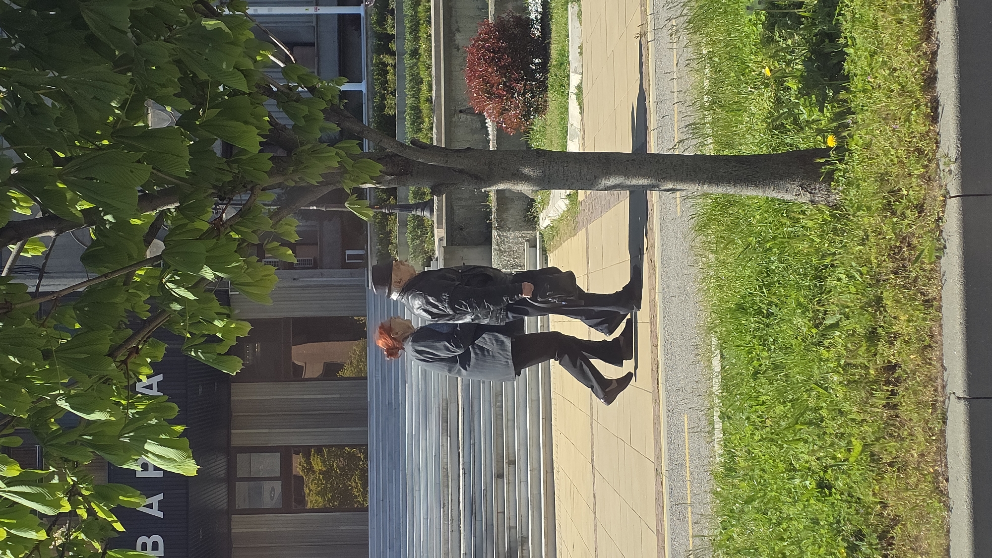 A medium-shot captures a scene on a sunlit sidewalk in Varna, Bulgaria. An elderly couple is walking: the woman has red hair, wears a dark jacket and pants. She leads slightly ahead. The man has grey hair and wears a dark hat and jacket. They are on a paved sidewalk adjacent to a patch of green grass, with a tree to the left and a building behind them. The building has reflective windows and stairs. A sign with white Cyrillic letters is visible on the building.