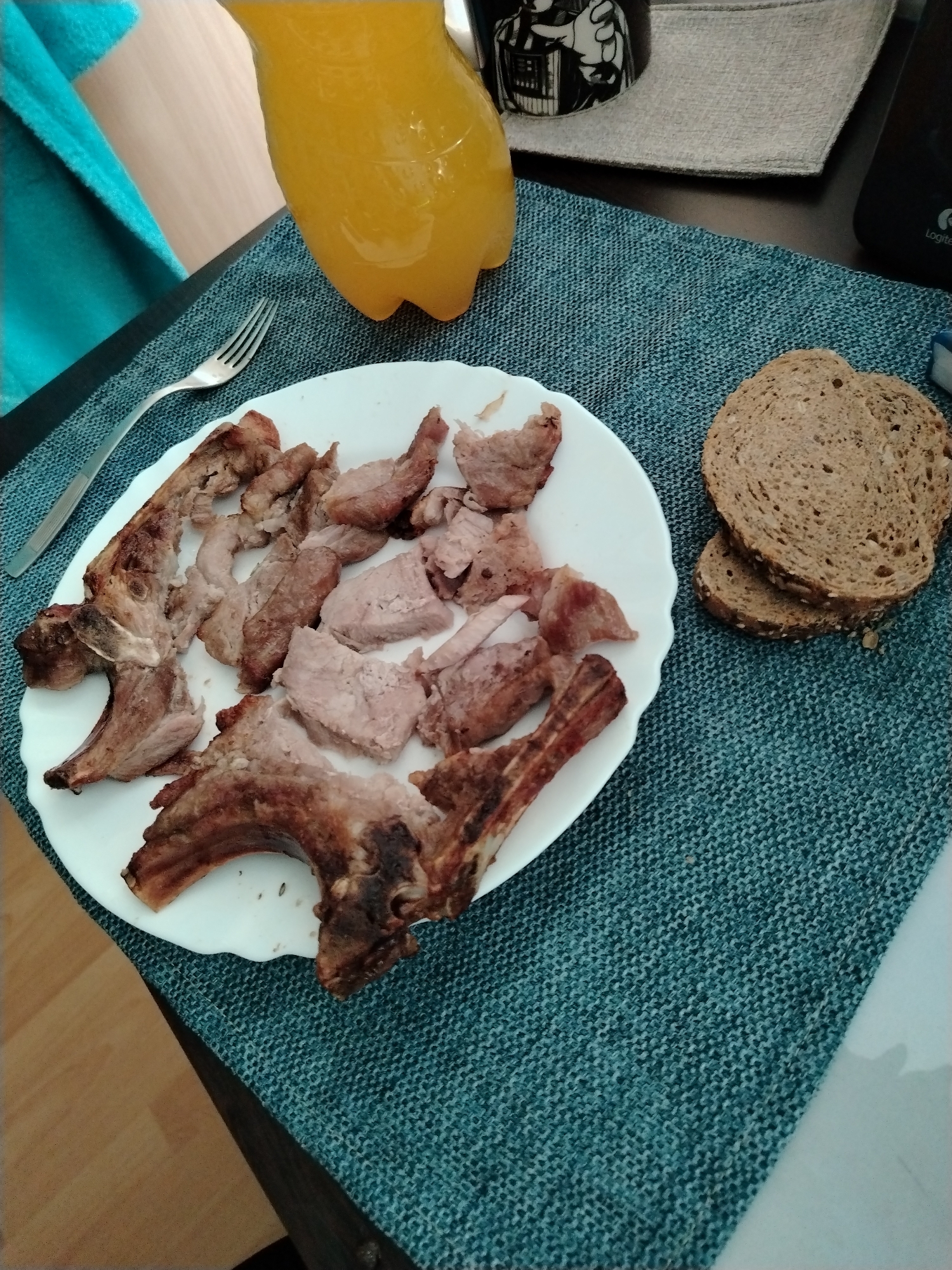 In the city of Silistra, Bulgaria, a meal is displayed on a dark table covered by a teal-colored, tightly woven placemat or table runner. A white plate with a subtly scalloped edge occupies the left side of the placemat, containing numerous pieces of cooked meat, some of which are identifiable as pork ribs with distinct bone structures, while others are boneless cuts. The meat exhibits varying degrees of doneness, from lighter pinkish-brown to well-browned and slightly charred edges. To the left of the plate, a silver-colored metal fork rests on the placemat. On the right side of the placemat, three slices of dark, seeded bread are stacked. In the upper left background, a clear plastic bottle filled with a yellow-orange liquid, likely a beverage such as orange juice, stands upright. Further back and to the right, a dark mug featuring a white graphic, possibly depicting Darth Vader, is partially visible, resting on a light grey fabric coaster. A dark electronic device, possibly a speaker or part of a computer, bearing a "Logitech" logo, is visible in the far upper right background. A bright turquoise or light blue towel is draped over an unseen object in the far upper left. The table surface is dark brown, and a light wooden floor is visible in the lower left corner of the scene. No individuals are present in the image.