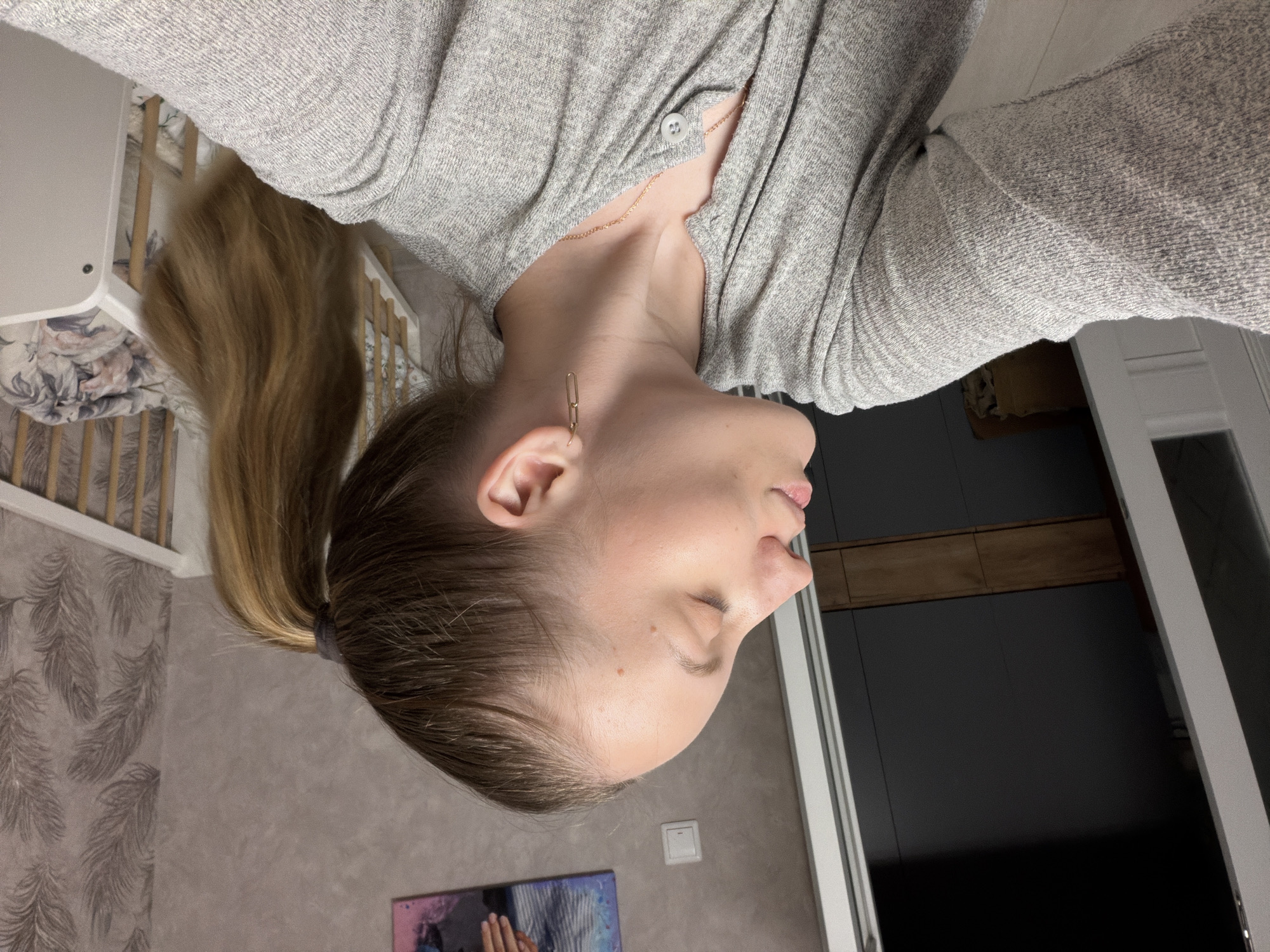 A woman with light skin and brown hair pulled back in a ponytail is depicted from the side. She is wearing a light gray long-sleeved shirt and gold earrings. A gold necklace is visible at her neck. Her eyes are closed. The background includes a wall with a pattern, a crib, a painting, and a doorway with a dark interior. The location is not discernable from the image alone.