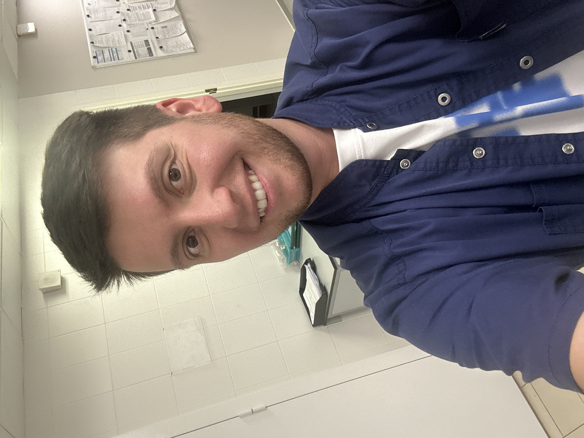 A man is taking a selfie in what appears to be a tiled room, possibly a laboratory or medical facility. He is wearing a dark blue scrub top over a white t-shirt with blue markings. The man has dark hair and is smiling, showing his teeth. He has a short beard. In the background, there is a bulletin board with papers pinned to it, a desk with papers in a tray, and a basket with plastic bags and other items. The walls are covered in white tiles, and there is a white door on the left side of the image. The lighting is bright, suggesting artificial light sources. There is no indication of the location being in Moscow, Russia.