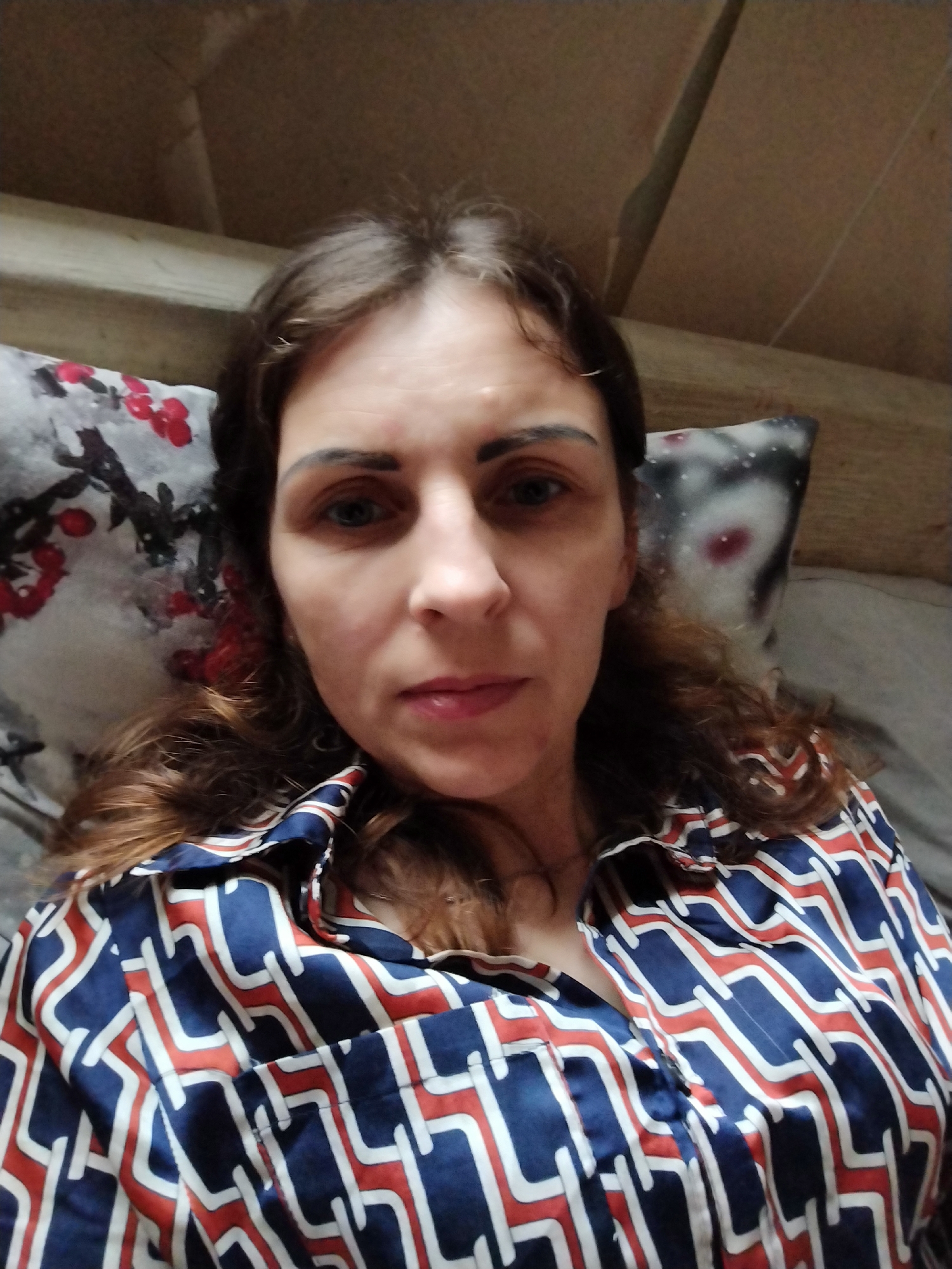 The image shows a selfie of a woman with brown hair, blue eyes, and fair skin. She is wearing a collared shirt with a blue, white, and red geometric pattern. The woman is lying down, and behind her, there are two pillows. One pillow has a floral pattern with red flowers. The background appears to be an indoor setting with wooden elements visible. The lighting is dim. The specific location in Vinnytsia, Ukraine, is not discernible from the image itself.