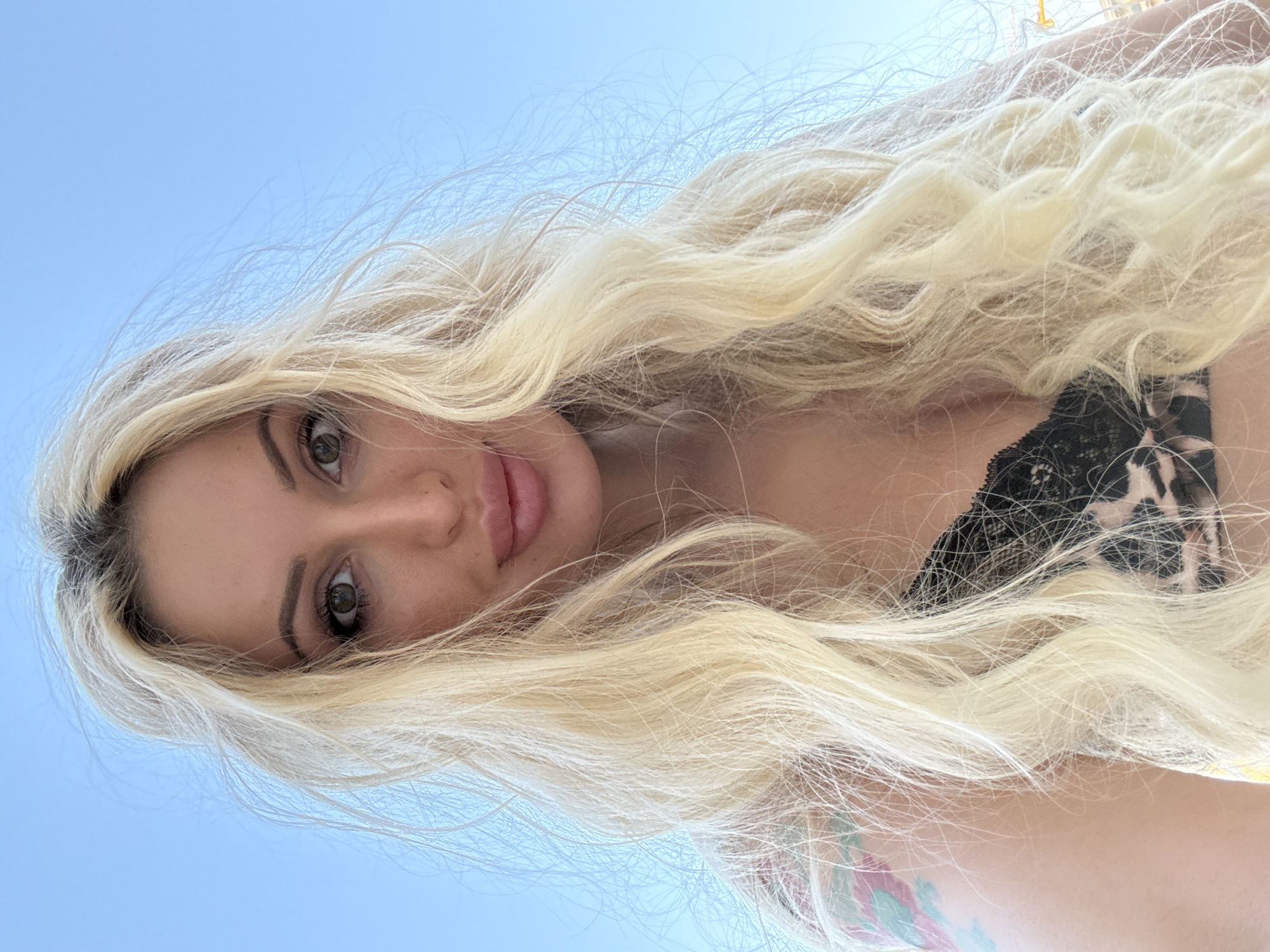 A woman with long blonde hair and green eyes is pictured from the chest up. She is wearing a black lace bra with a leopard print design. She has a tattoo on her left shoulder that appears to be abstract with pink, blue, and green colors. The background is a clear blue sky. The lighting suggests it is daytime and sunny. There are no other people or objects clearly visible in the frame.
