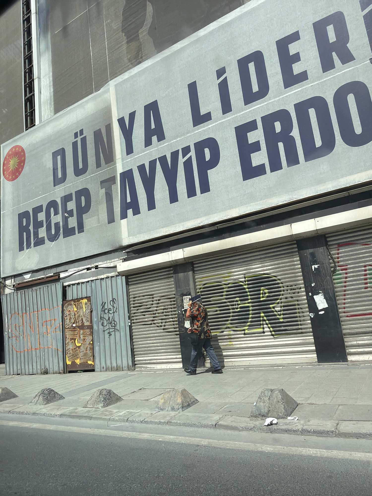The image depicts a street scene in Bahçelievler, Turkey, dominated by a large banner on a building under construction. The banner, predominantly white with dark blue text, reads "DÜNYA LİDERİ RECEP TAYYİP ERDOĞAN" in large, bold lettering. To the left of the text is a circular emblem with a star and crescent, likely a Turkish national symbol. The banner extends across the facade, partially obscuring the building's structure.
Below the banner, the ground level features several metal roll-up doors, some of which are graffitied. One door on the left has a distinct yellow and brown spray-painted design, while another door in the center has green and yellow graffiti. To the far right, a black metallic structure is visible, possibly an electrical box or part of the building's facade.
A single individual, dressed in a patterned shirt and blue jeans, is walking on the sidewalk in front of the roll-up doors. The person appears to be holding something to their ear, suggesting they are on the phone. Several concrete bollards, pyramid-shaped and conical, are lined up along the edge of the sidewalk, separating it from the road. There is some debris on the sidewalk, including a crumpled white piece of paper. The road in the foreground is asphalt. The overall impression is a busy urban environment with elements of construction, advertising, and everyday street life.