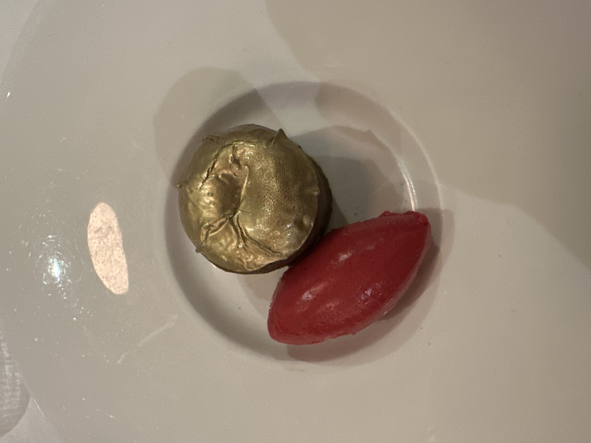 A top-down shot depicts a dessert on a white plate. The dessert consists of two distinct elements: a gold-colored, dome-shaped item that appears textured and possibly cracked on the surface, and a red, oblong, smooth-textured element, potentially a sorbet or fruit puree. The dessert is presented in the center of the plate, within a slightly recessed area. The lighting is diffused, and there is a faint reflection visible on the plate's surface. The scene lacks any additional context to ascertain the location or any human presence.