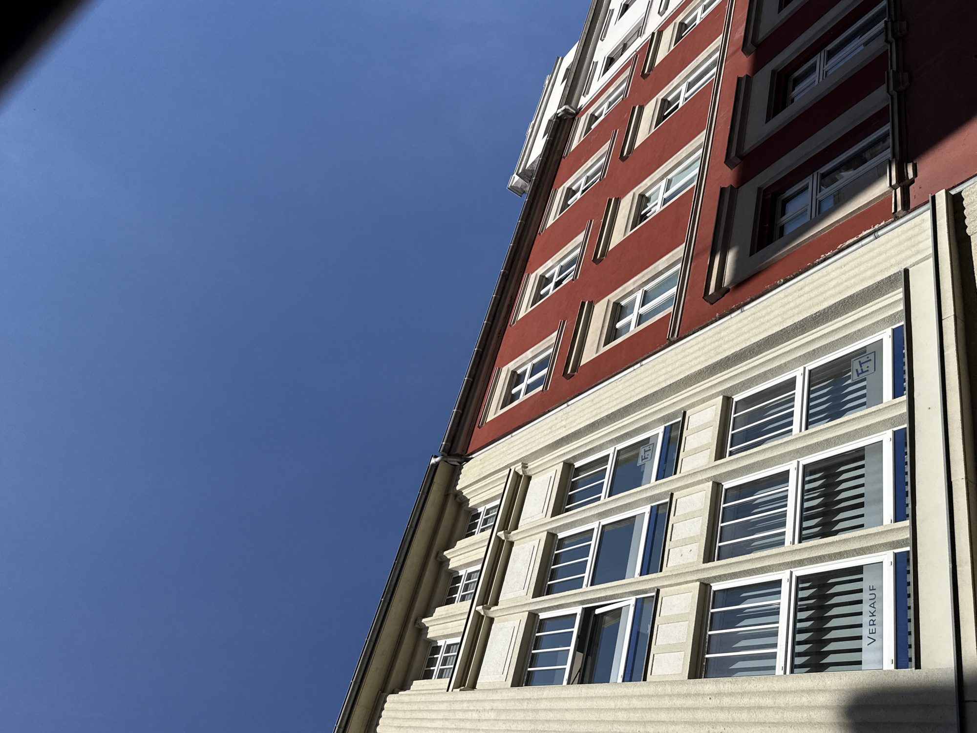 The image shows a view of buildings in the city of Görlitz, Germany, looking up at a clear blue sky. The buildings are adjacent to each other, with the left building being a pale beige color with classical architectural details like fluted columns. This building has several rows of windows with white frames. One of the lower windows has a sign that reads "VERKAUF," which means "for sale" in German, and a logo with the letters "F.T.".
The building to the right is painted a vibrant red and also features multiple rows of windows with white frames. The red building has a more modern facade with horizontal decorative elements above some of the windows. The architecture suggests an urban setting with a mix of older and possibly renovated structures. The angle of the shot emphasizes the vertical lines of the buildings against the expansive sky.