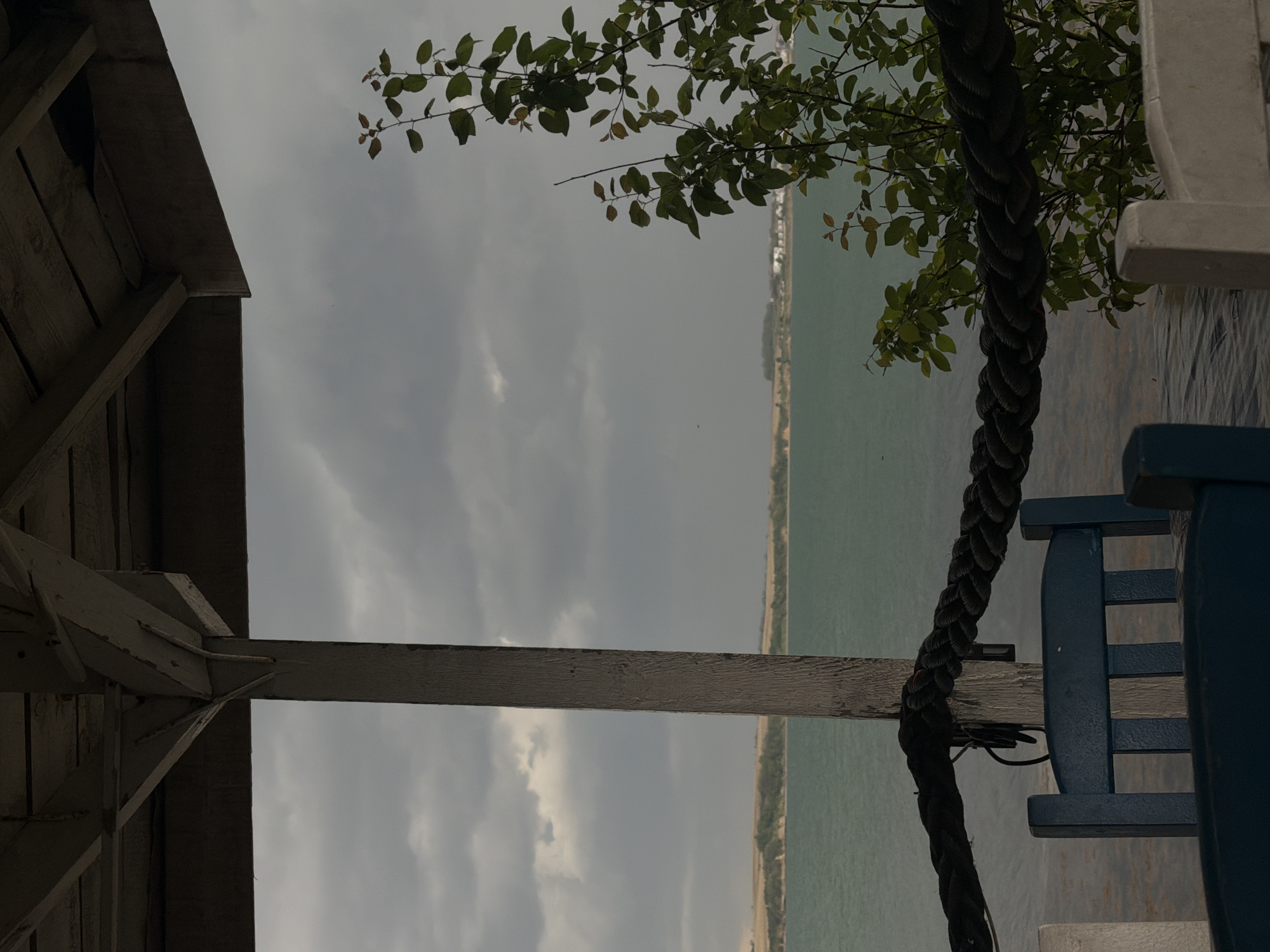 A still, rotated view presents an outdoor scene of a body of water, possibly a bay or ocean, stretching towards a distant, low-lying landmass beneath an overcast sky. The foreground contains elements of constructed spaces, notably a wooden structure (likely a building or shelter) framing the left side, featuring weathered, light-colored wood. A dark rope hangs vertically, adding texture to the right. Part of a blue object, which appears to be a wooden chair, is visible as well. Green foliage is present near the top center. The environment overall is subdued, suggesting a calm and possibly coastal location. The location in the city of Unknown, Unknown is uncertain.