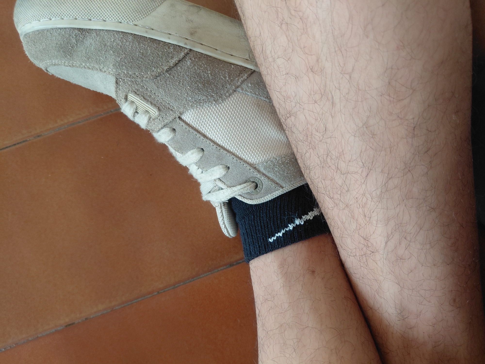 A close-up, medium shot shows a person's legs and foot wearing a tan suede and mesh sneaker. The shoe is laced with white laces. The person is wearing a dark sock with a white zig-zag pattern. The legs are covered in light to medium hair. The background is a tiled floor with an orange-brown hue. There is no indication of the location being Santa Ana, Venezuela.