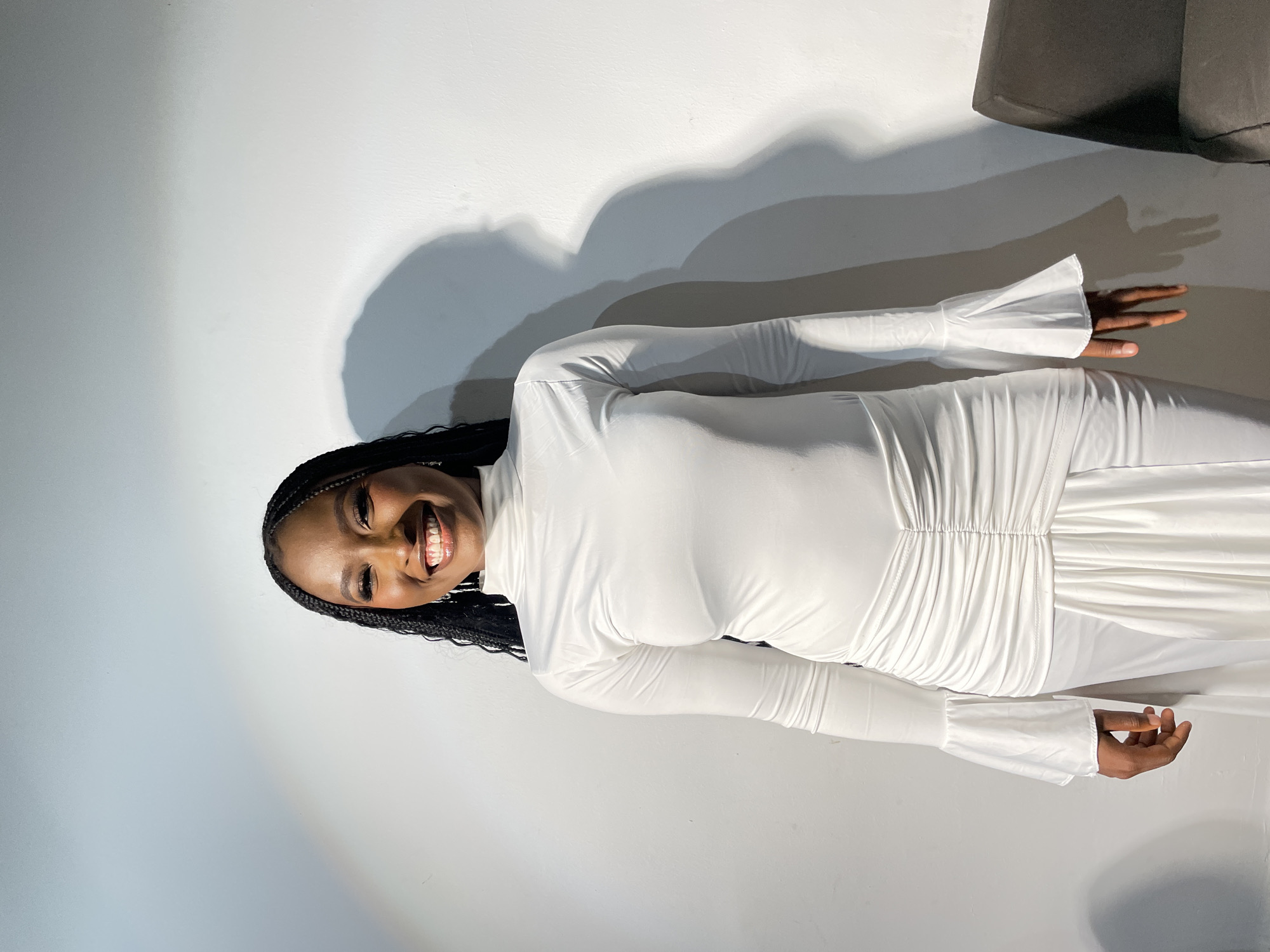 A woman with dark skin and braided hair is standing indoors against a white wall. She is wearing a long-sleeved, white dress. The dress has gathered detailing around the waist and flared sleeves. A gray chair is visible in the upper right corner of the frame. The lighting in the room creates a shadow on the wall behind the woman. The location is not evident in the image and I cannot provide any location information.