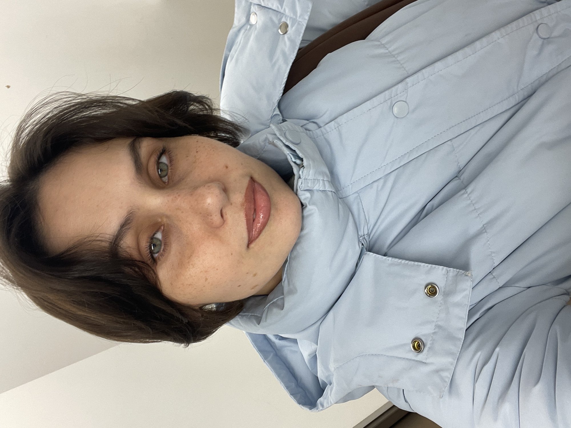 A woman with dark brown hair, fair skin with freckles, and green eyes is looking at the camera. She is wearing a light blue puffy jacket. The image is a selfie or close-up shot, with the woman's face and upper torso prominently displayed. The background appears to be a plain white wall. There is no indication in the image that it was taken in Izhevsk, Russia, or any specific city.