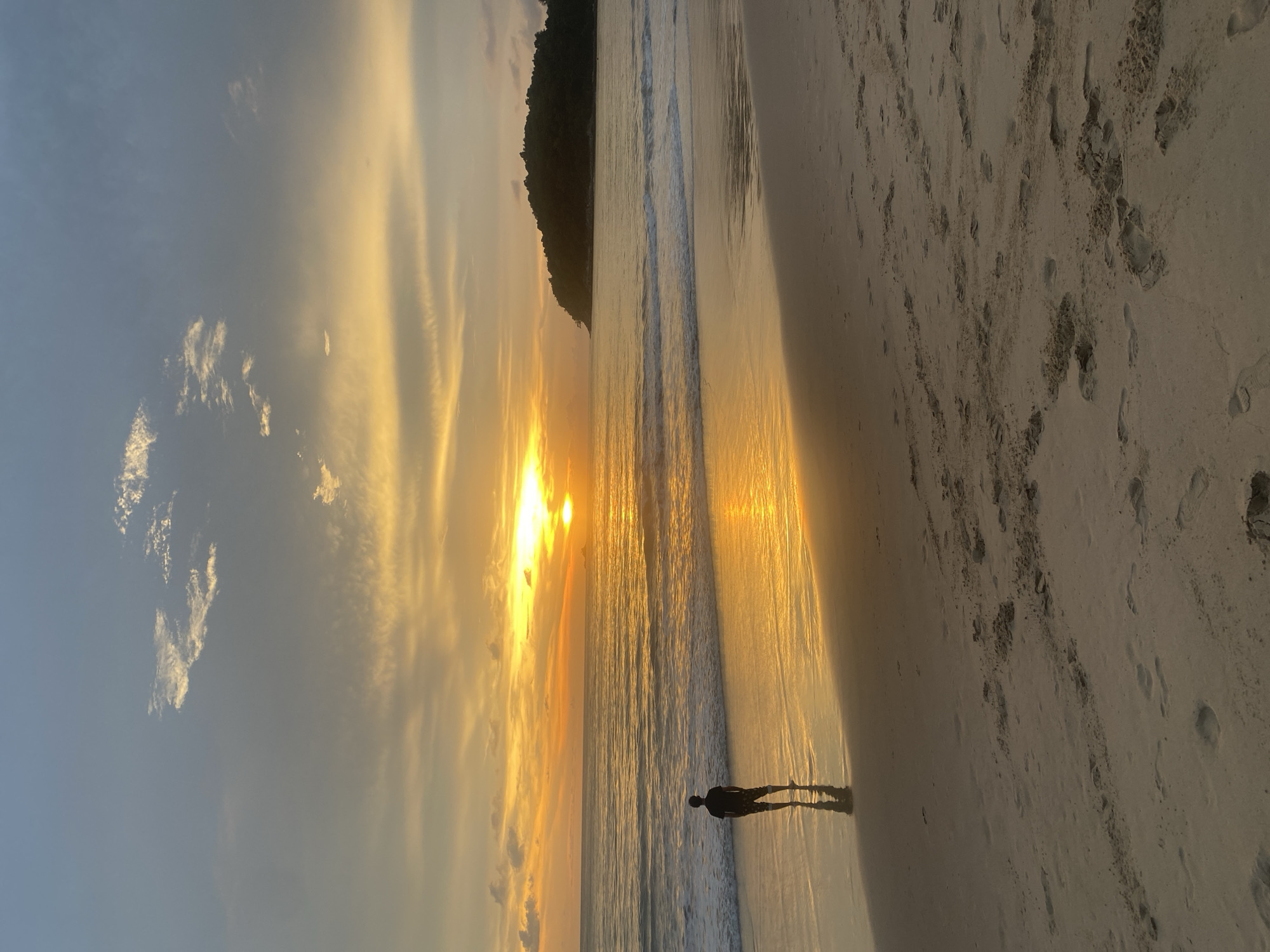A vertical, eye-level photograph captures a beach scene at sunset, likely in Choeng Thale, Thailand, given the presence of tropical foliage in the upper frame. The setting sun dominates the horizon, casting a golden glow across the water and sand. A silhouetted figure stands at the edge of the water, casting a long shadow on the wet sand. Footprints are visible on the dry sand. A dark land mass or small mountain is present on the horizon. The sky is filled with golden-tinged clouds. The scene is calm and tranquil.