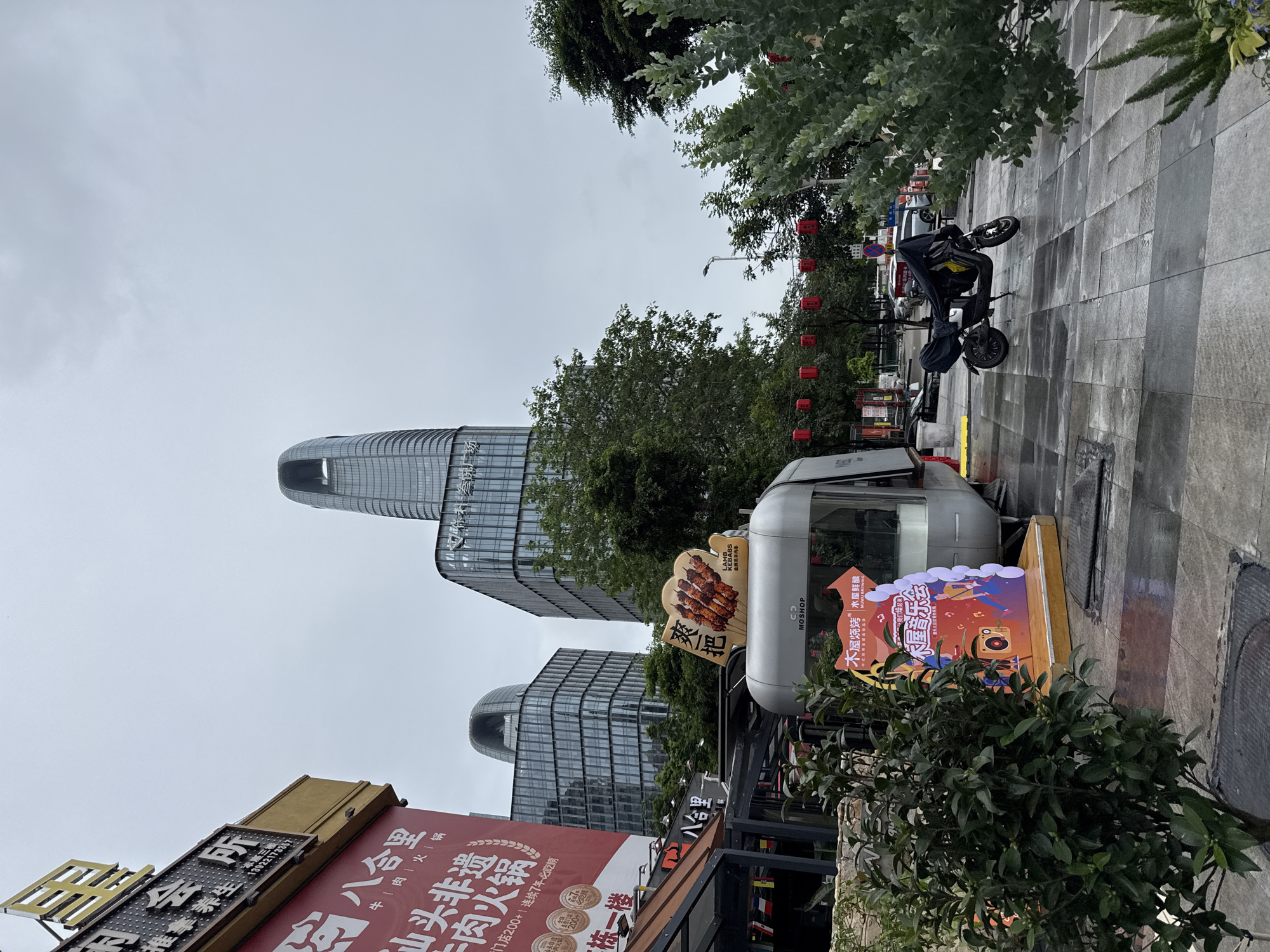 The image captures a street scene in Haizhu District, China, under an overcast sky. In the foreground, a modern, silver food truck or trailer is parked on a wet, tiled pedestrian walkway. A black electric scooter is parked next to the food truck, partially covered with a black tarp. To the left of the food truck, a green shrub with dense foliage is visible. A tall, red banner advertising "八合里牛一肉火锅" (Baheli Beef and Mutton Hotpot) hangs on the side of a building. Further back, the scene is dominated by contemporary glass-fronted skyscrapers, one of which has a distinctive curved, open top. Red lanterns are strung across the street in the mid-ground. The overall environment suggests an urban commercial area.