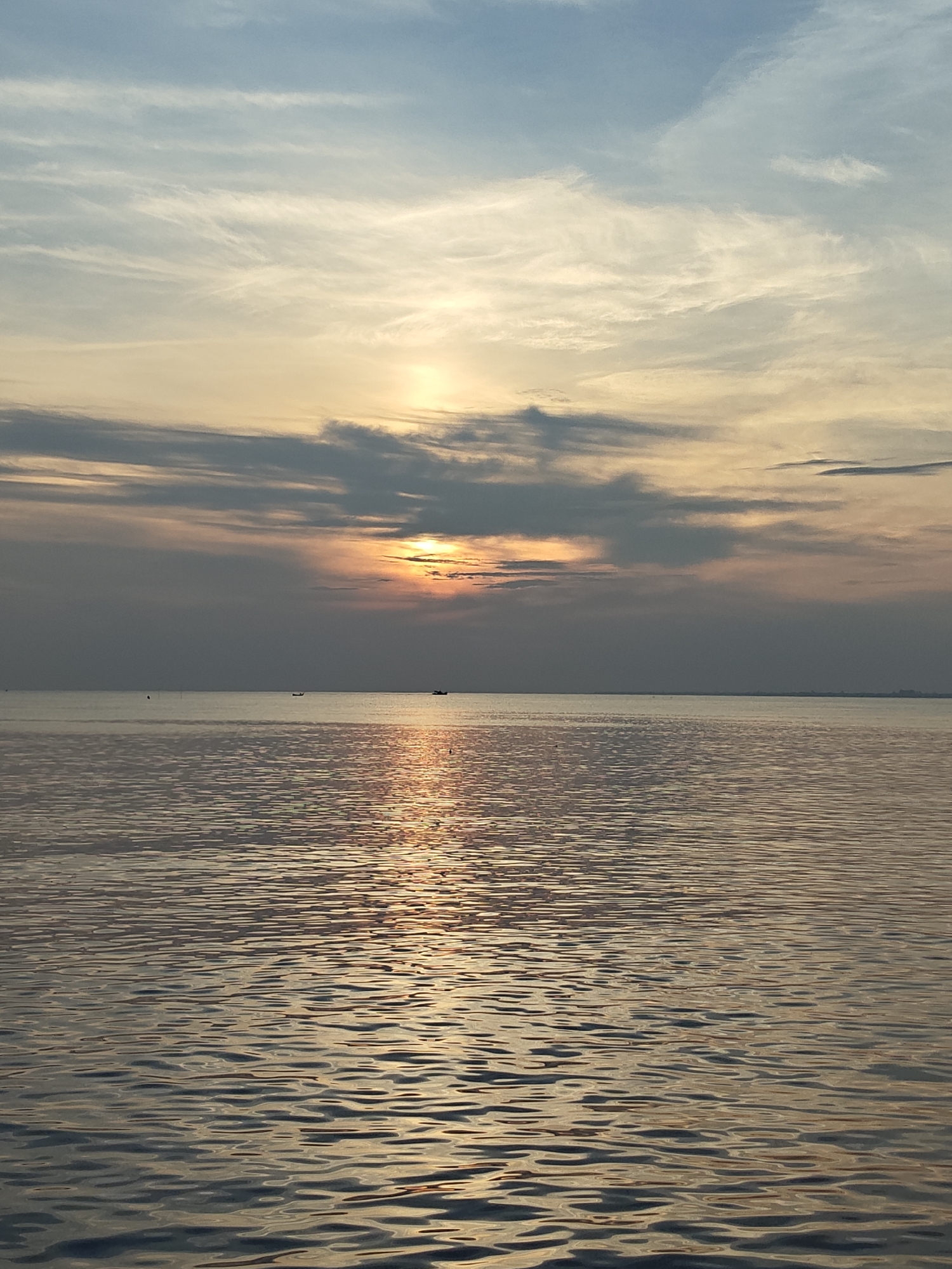 The image depicts a serene ocean scene at sunset, with the sun partially obscured by a band of clouds. The water is calm, reflecting the golden light of the setting sun in a shimmering path toward the viewer. In the distance, the horizon is visible, with faint outlines of land. There are three small boats on the water, appearing as tiny silhouettes. The sky above the clouds features a mix of blue and golden hues, characteristic of twilight. Based on the prompt, this image likely depicts an ocean view in Lhokseumawe, Indonesia, characterized by a flat coastline.