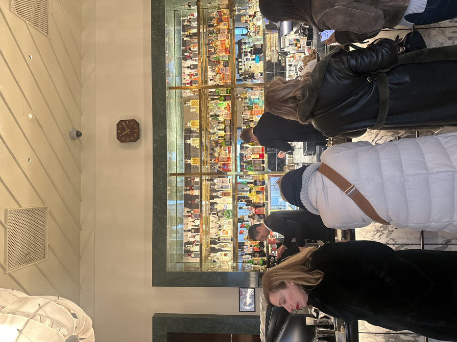 The scene depicts the interior of Camparino in Galleria, a prominent bar located within the Galleria Vittorio Emanuele II in Milan, Italy. The vantage point is from the customer side of a busy bar, looking towards the main service counter and liquor display.
In the background, a large, well-stocked liquor display spans the width of the image, featuring numerous bottles of various spirits arranged on multiple tiers of brass-framed glass shelves, illuminated from behind. This display is set within a dark green marble wall cladding. Centered above the liquor display on a white wall, an octagonal wooden clock with gold hour markers and hands indicates the time. The ceiling is white, with linear recessed lighting, two square perforated metallic ventilation grilles, and a dome-shaped security camera visible. A large, modern light fixture with white, draped fabric elements is partially visible in the upper left corner.
In the midground, behind the white marble bar counter, two male bartenders in dark suits are actively working. On the left, a bartender with glasses is attending to an espresso machine, preparing a beverage. On the right, another bartender is mixing a drink with a shaker. The bar counter itself is constructed from white marble with a distinctive vertical fluted design.
In the foreground, several patrons stand at the bar. On the far left, a woman with shoulder-length blonde hair, wearing a black fur-like coat, is visible in profile, looking to her left. Her right hand is near a small white cup on a black tray on the counter. To her right, a person with their back to the camera wears a white puffer jacket, a dark blue knitted hat, and has a brown shoulder bag strap visible. Further right, another person, also seen from behind, wears a dark, possibly leather or shearling-lined coat. On the far right, a partially visible person with dark hair holds a smartphone in their left hand, observing the screen.
The environment is brightly lit, suggesting an upscale establishment with a classic yet elegant design.