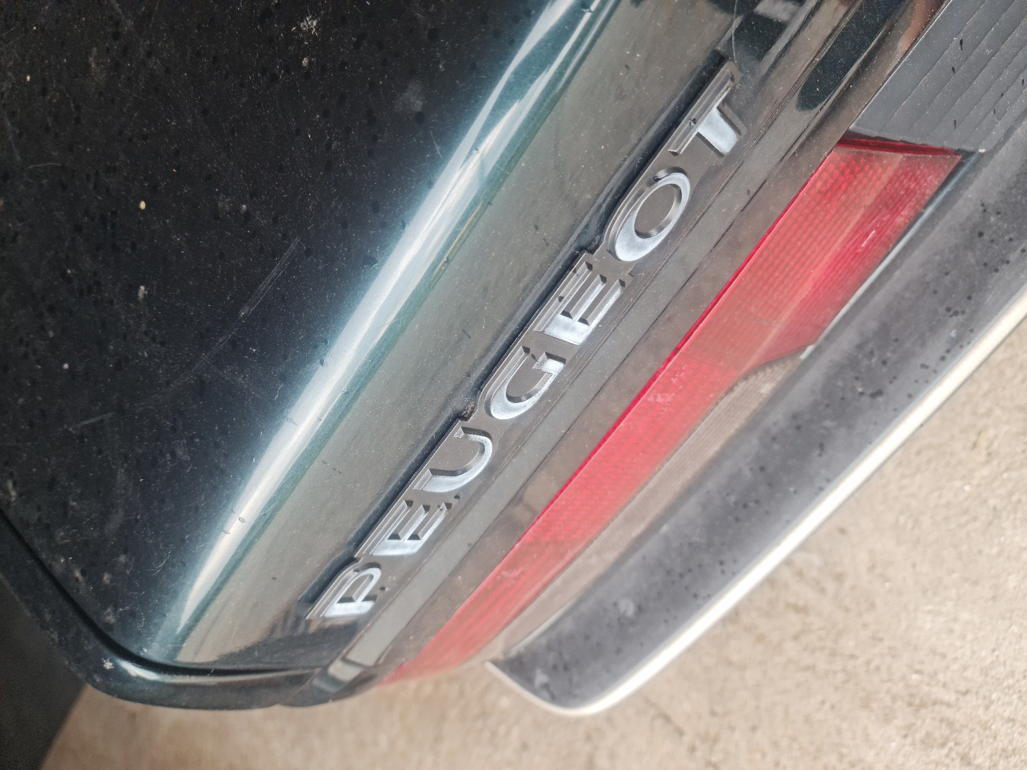 The image presents a close-up, angled view of the rear of a dark-colored car, likely situated outdoors in Buzovgrad, Bulgaria. The car's bodywork appears to be a very dark green or black, covered with a layer of dust, dirt, and minor surface scratches.
The most prominent feature is the silver, raised lettering on the car's rear panel, clearly spelling out "PEUGEOT". These letters have a metallic sheen, reflecting the ambient light. To the right of the lettering, a portion of a red, rectangular taillight or reflector is visible, also showing signs of dust accumulation. The bottom right edge of the frame reveals a light-colored ground surface, possibly concrete or asphalt, indicating the vehicle is parked on a paved area.
The scene depicts a stationary vehicle; no activity or people are present. The reflections and general brightness suggest it is daytime, with either an overcast sky or diffused sunlight. The car's dusty condition implies it has been exposed to outdoor elements.