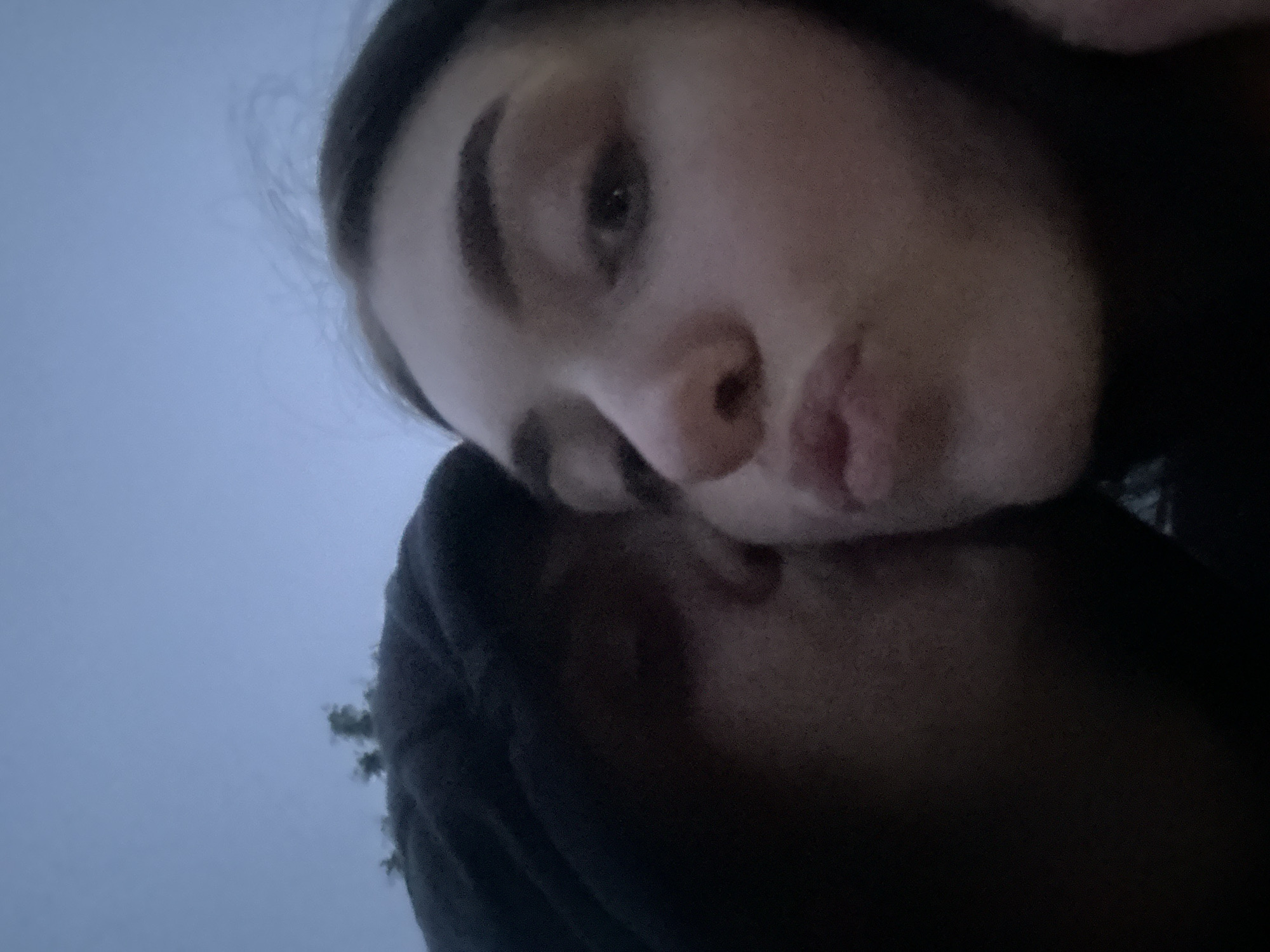 A low-angle, slightly blurred photograph shows two individuals, presumably a couple, in an embrace. The individual in the upper portion of the frame is lighter-skinned with dark hair and eyebrows. Their face is positioned towards the camera, with their lips pursed slightly. The second individual is partially obscured, with only a portion of their face visible resting against the first person's head. They are wearing a dark-colored hooded garment. The background appears to be an overcast sky, and it is impossible to determine if the photo was taken in Burgas, Bulgaria, or elsewhere.