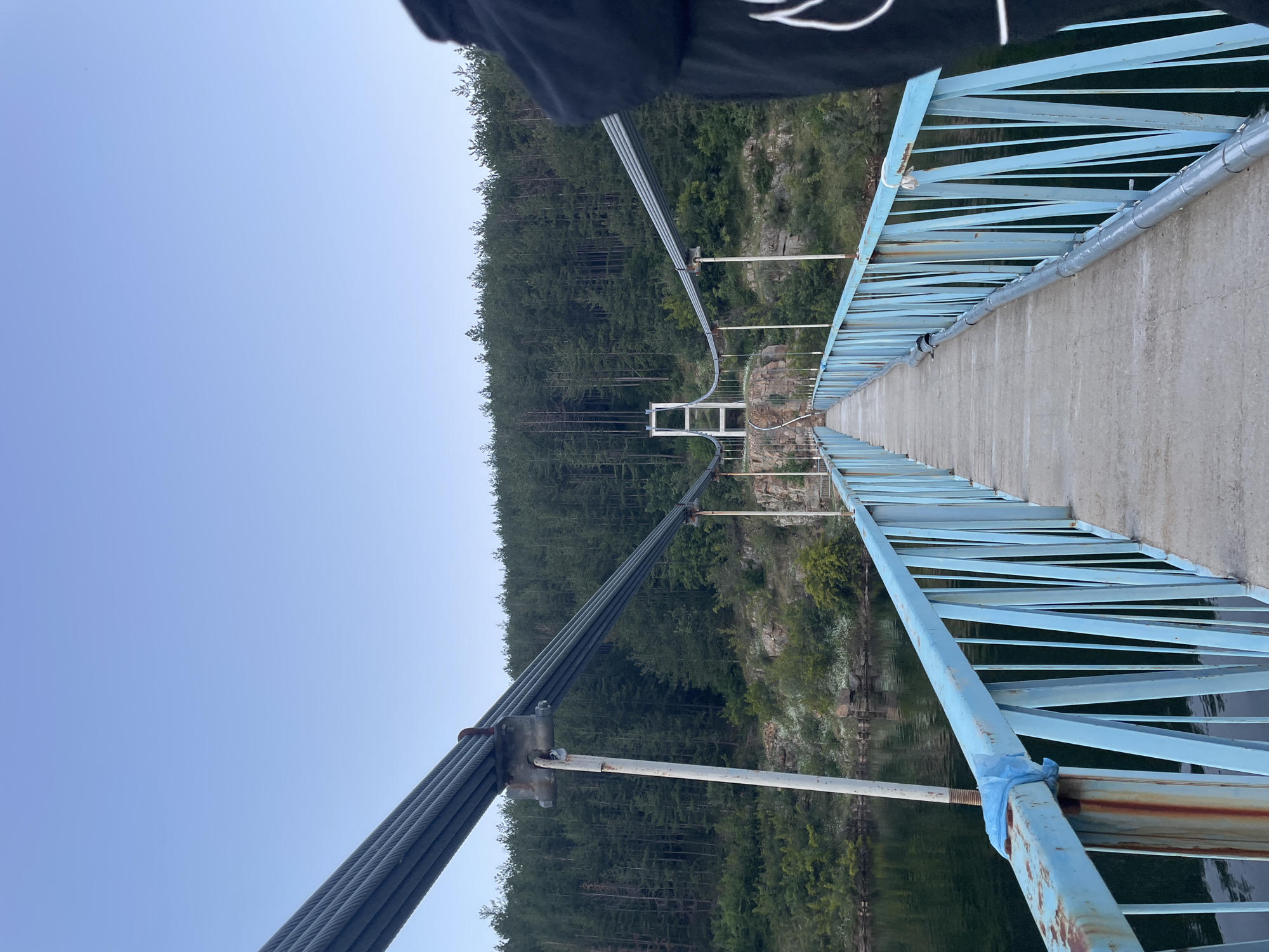 The image shows a suspension bridge, likely the Bridge over Yovkovtsi Reservoir, located near the town of Suhinvo, Bulgaria. The bridge has a concrete walkway with light blue metal railings. Thick steel cables stretch overhead, supporting the bridge. On either side of the bridge, the landscape features a calm body of water, rocky terrain, and a dense forest of pine trees. The sky is clear and blue, suggesting daytime. The photograph is taken from the perspective of someone standing on the bridge, looking towards the far end. A person's arm, wearing a black hoodie with a white design, is visible on the right side of the frame, indicating the photographer's presence. The overall scene conveys a sense of natural beauty and a recreational area.
