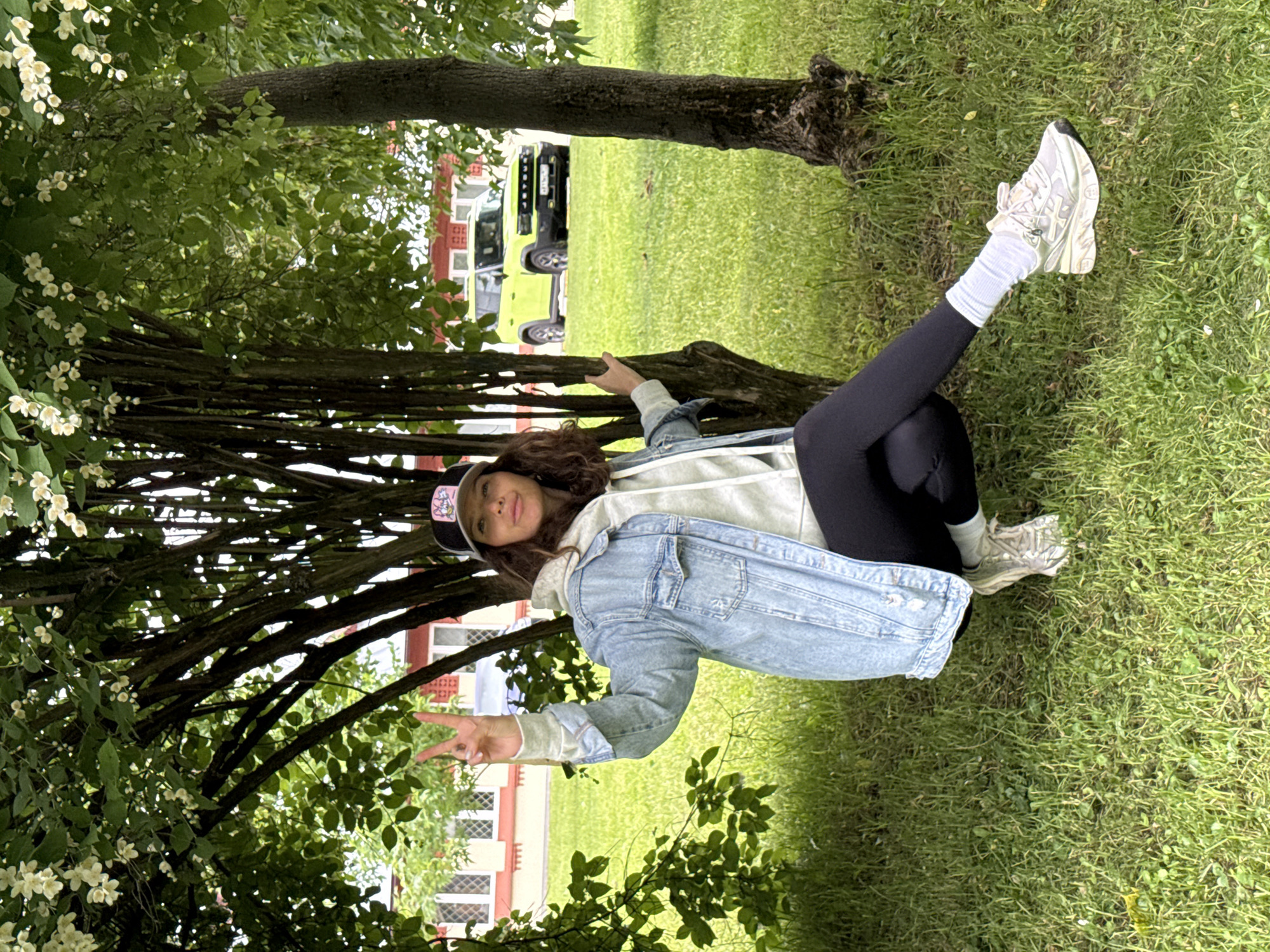 A woman is squatting on the grass next to a tree, making a peace sign with her left hand. She is wearing a denim jacket, a grey hoodie, black leggings, white socks, and light-colored sneakers. A bright yellow SUV is parked in the background. The scene appears to be in a park or green space with buildings visible behind the vehicle.