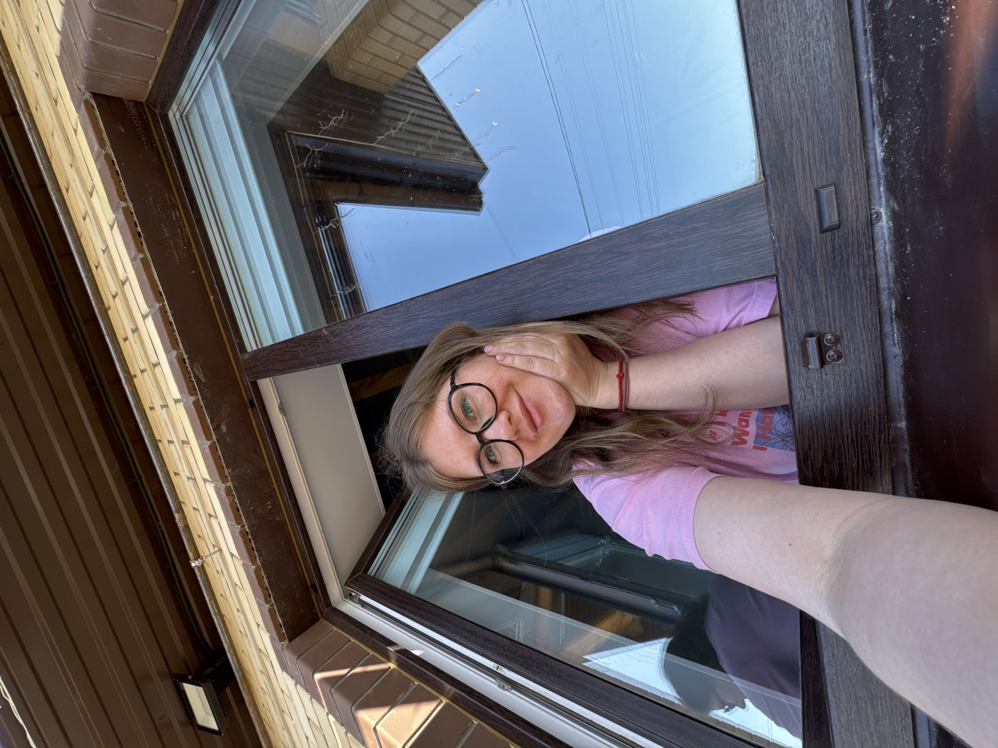 A woman with glasses is looking out of an open window. The window frame is dark brown. She is wearing a pink shirt and has light brown hair. The wall around the window is made of light brown and dark brown siding. The location is indicated as Gornyi (Dvurechenskoe S/P), Russia. The sky is visible through the window, appearing light blue.