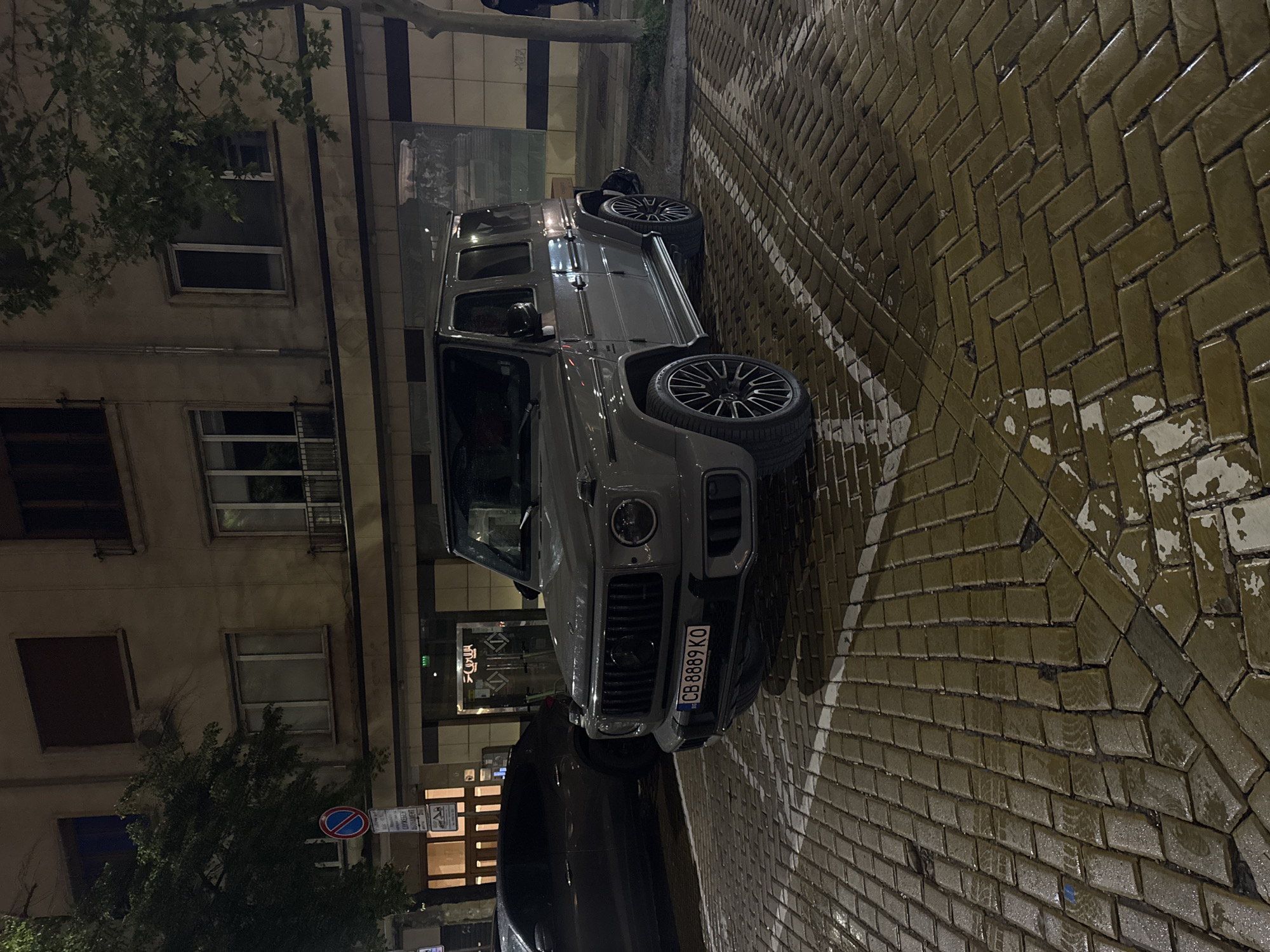 A gray Mercedes-Benz G-Class SUV is parked on a street in Sofia, Bulgaria. The license plate reads "CB 8889 KO." The street is paved with light brown, rectangular bricks in a herringbone pattern. A white painted line designates a parking space. A multi-story building is visible in the background, with windows and a business storefront displaying Cyrillic script. Street signs and trees are also visible in the foreground. The lighting suggests it is nighttime or dusk.