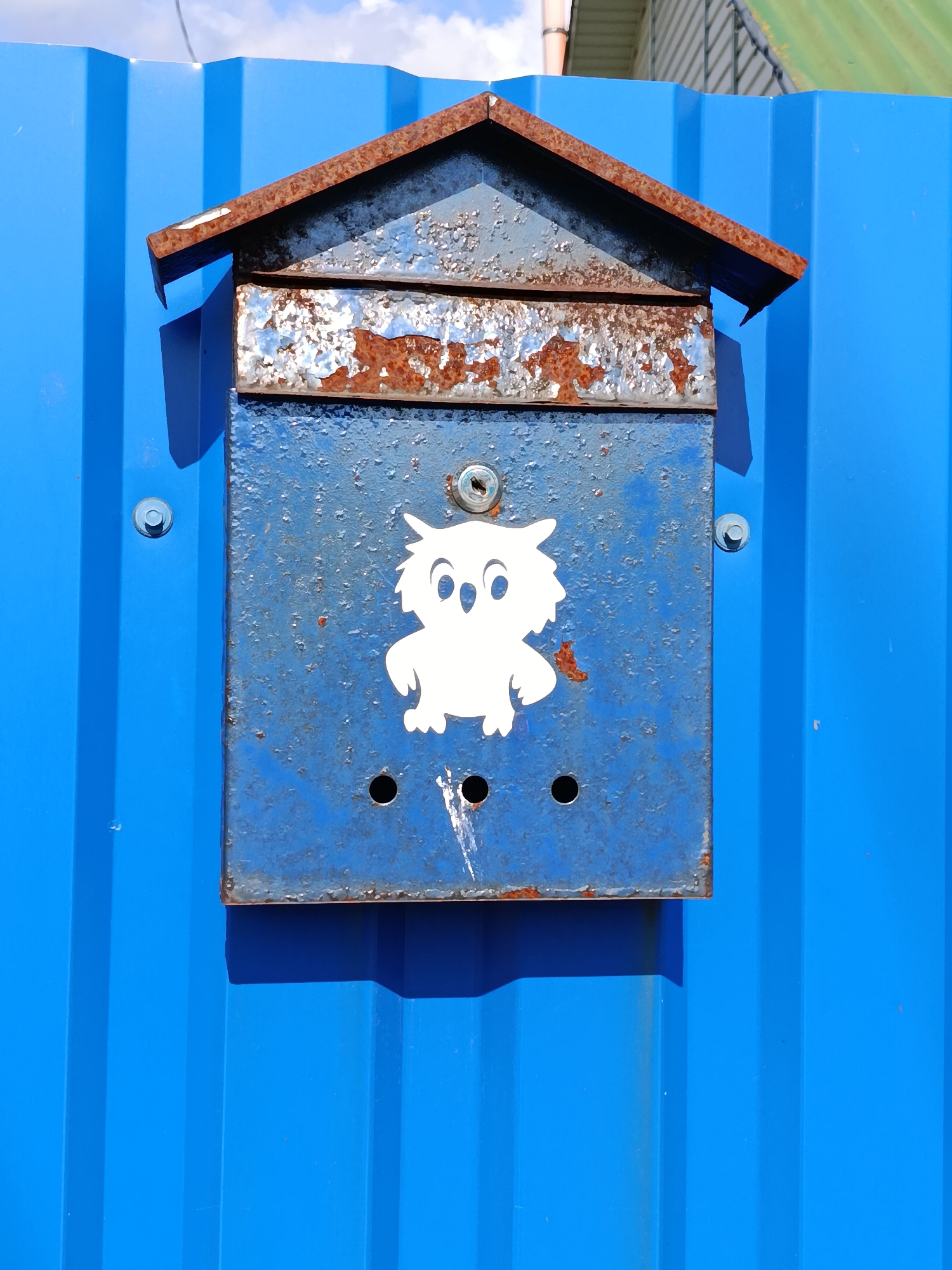 The image shows a weathered blue metal mailbox attached to a bright blue corrugated metal fence. The mailbox has a rustic, house-shaped roof made of rusted metal. A white sticker of a cartoon owl is affixed to the front of the mailbox. Below the owl sticker, there are three small holes in the mailbox. The mailbox is mounted to the fence with two visible bolts. The scene is outdoors, with a hint of sky and greenery visible at the top of the frame. The lighting suggests it is daytime. The location is specified as Смілавіцкі сельскі Савет, Belarus.