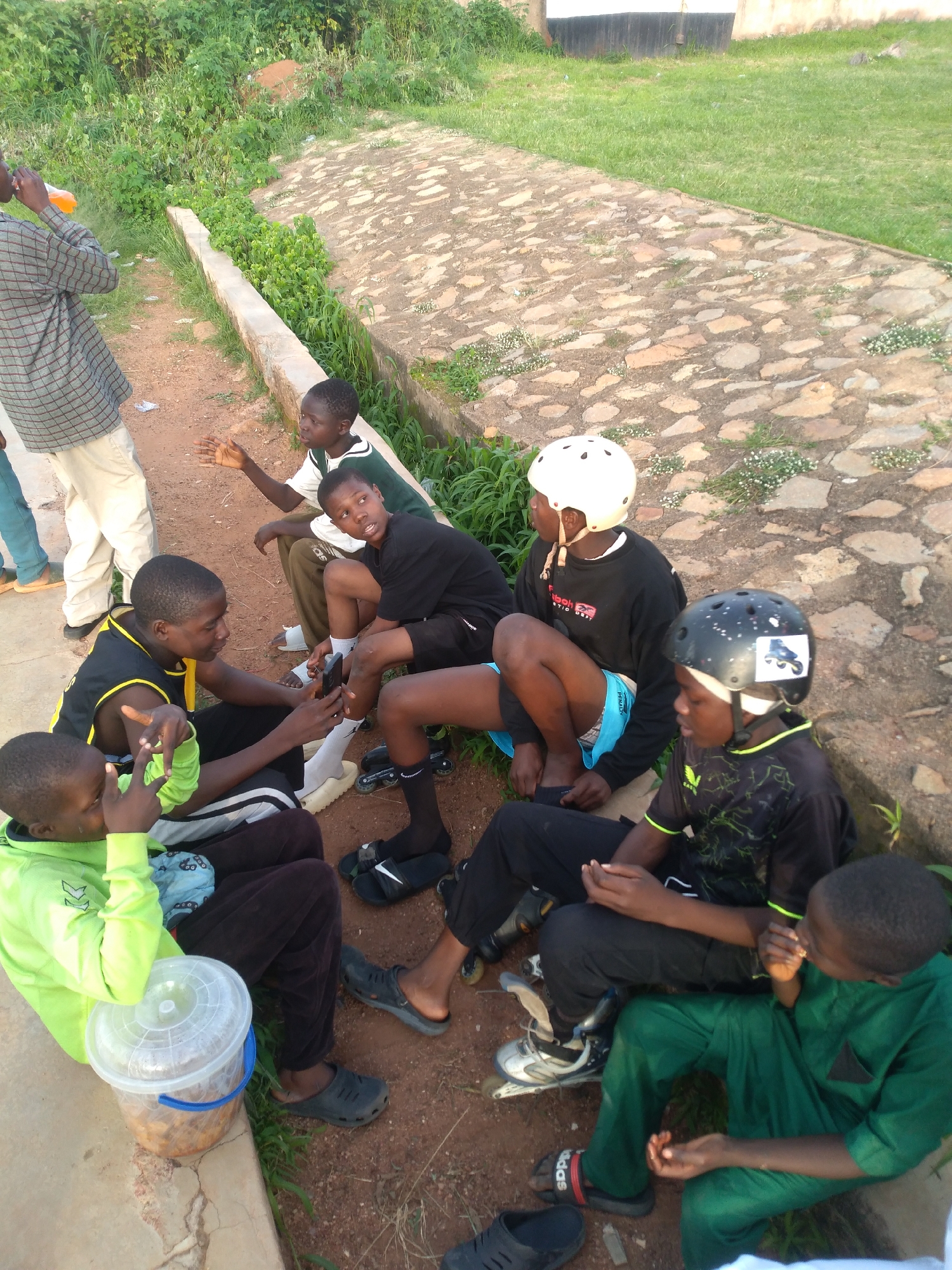 In Gura Top, Nigeria, a group of young boys are gathered outdoors. They are sitting on the ground and a low wall, some wearing protective helmets, suggesting they might be involved in activities like rollerblading or skateboarding. The environment appears to be a somewhat unpaved area next to a grassy embankment and a stone pathway. One boy is seen drinking from a bottle of orange liquid, and another is looking at a mobile phone. A transparent plastic bucket containing food is visible near one of the boys. The overall scene depicts a casual, relaxed gathering of young individuals.