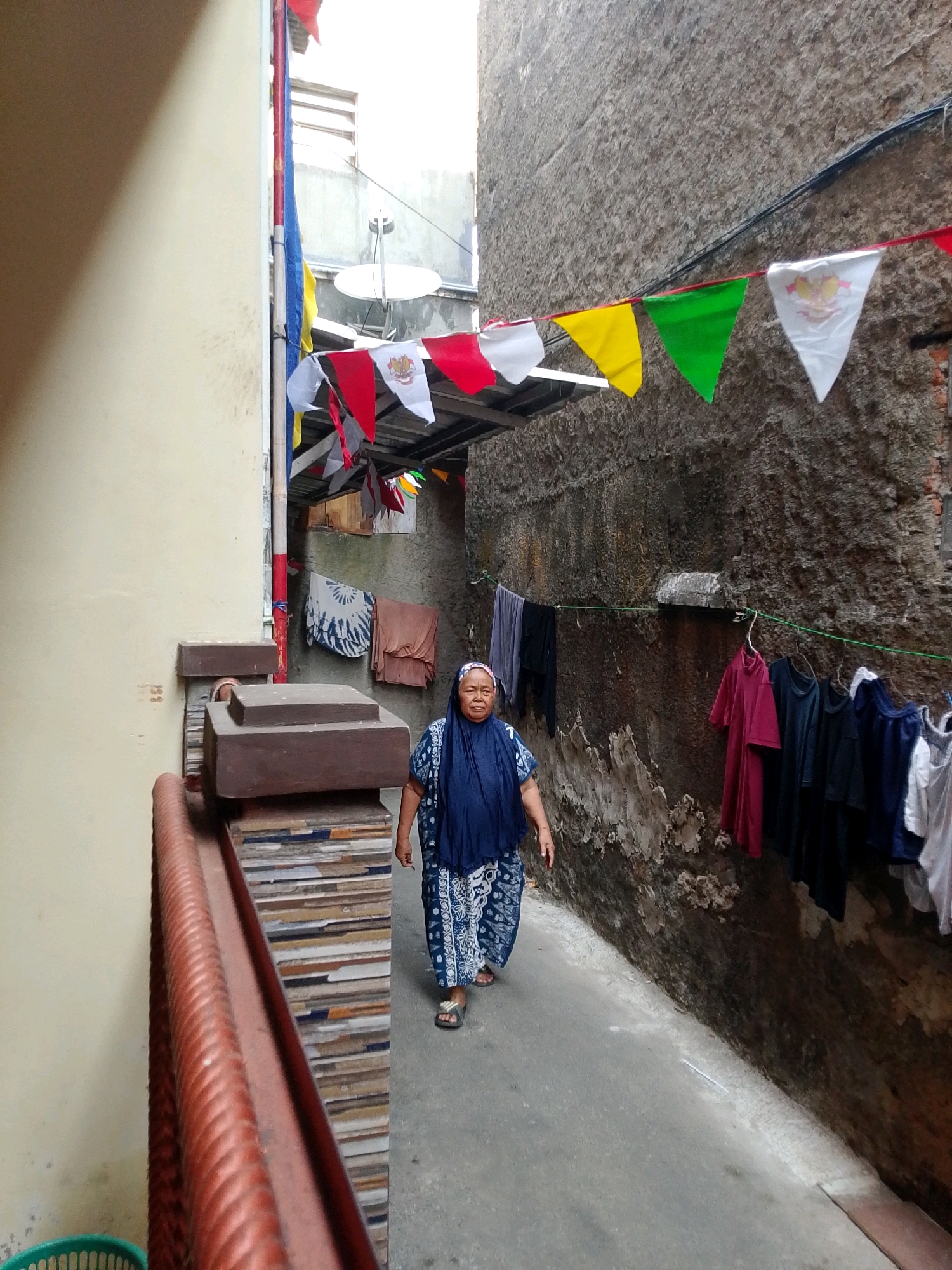 An elderly Indonesian woman wearing a dark blue headscarf and a blue and white patterned dress walks down a narrow alley in East Jakarta. The alley is flanked by a light-colored building on the left and a textured, dark grey stone wall on the right. Numerous colorful triangular flags are strung across the alley overhead, including red, white, yellow, and green flags. Clothes hang on a clothesline attached to the stone wall, with garments in various colors and styles, including a pink shirt, dark blue shirts, and white shirts. The ground is paved with concrete. In the foreground on the left, a red railing and a section of a tiled structure are visible.