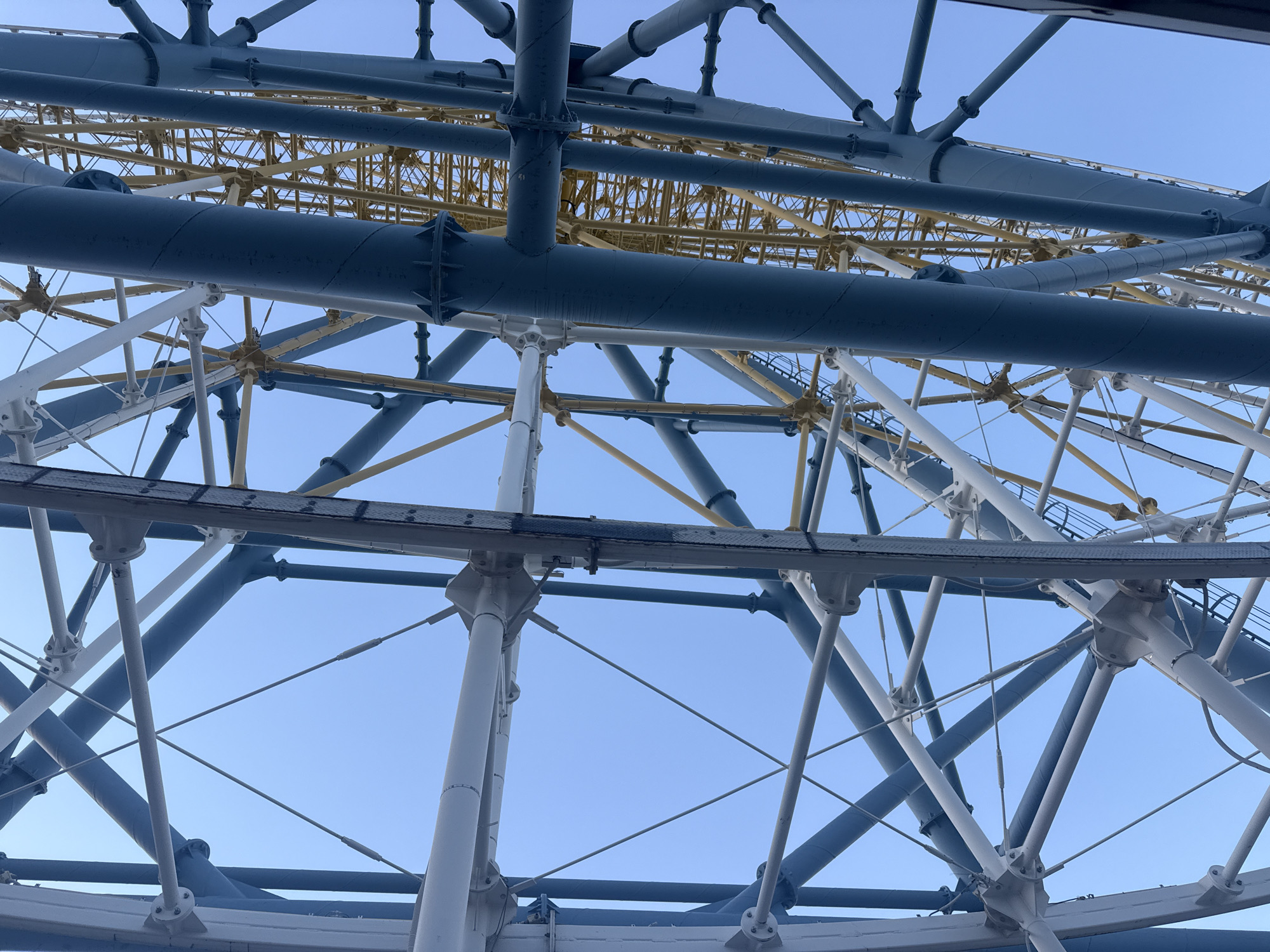 The image shows a section of the Tbilisi Ferris Wheel against a light blue sky. The Ferris wheel structure is comprised of intersecting beams, some painted in white, others in blue and yellow.