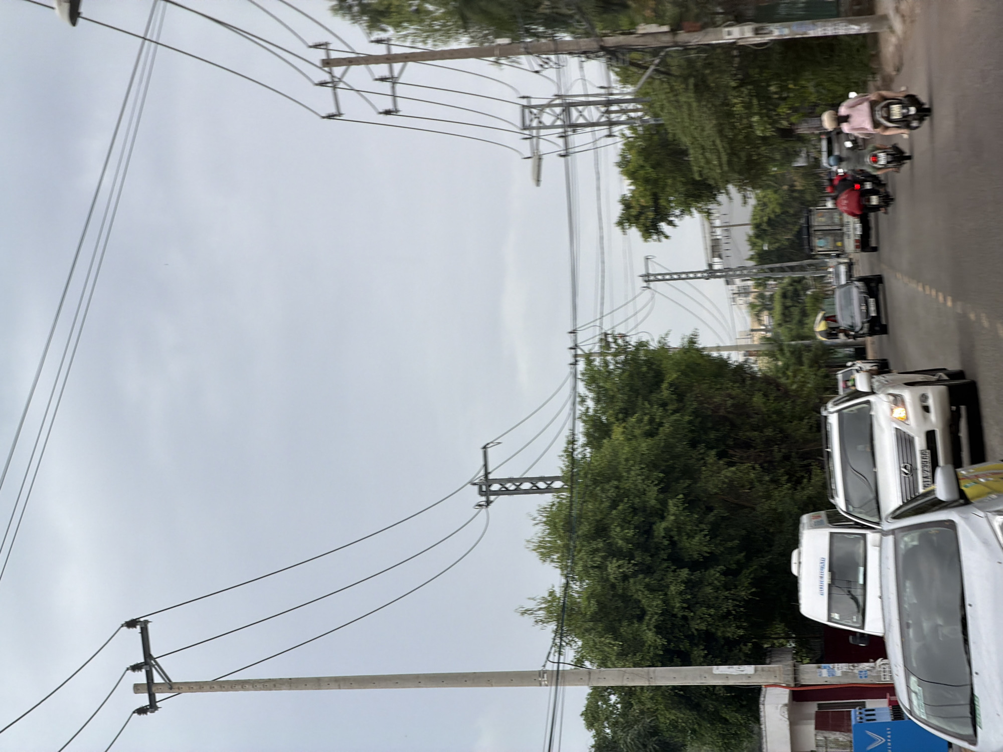 This image depicts a street scene in Nha Trang, Vietnam, characterized by a cloudy sky and overhead power lines. The road is congested with vehicles, including cars, vans, and motorcycles. In the foreground, a silver car is visible, partially obscuring a white van and a white Lexus SUV. The Lexus SUV has its headlights on and a license plate reading "61A-739.66". Further down the road, several motorcycles are seen, some carrying multiple passengers. One motorcycle in the middle ground has a rider in a red jacket, and another has a rider wearing a pink shirt and a helmet. In the background, lush green trees line the street, and further beyond, buildings and more power line infrastructure are present. The overall atmosphere suggests a typical day of traffic in an urban environment.