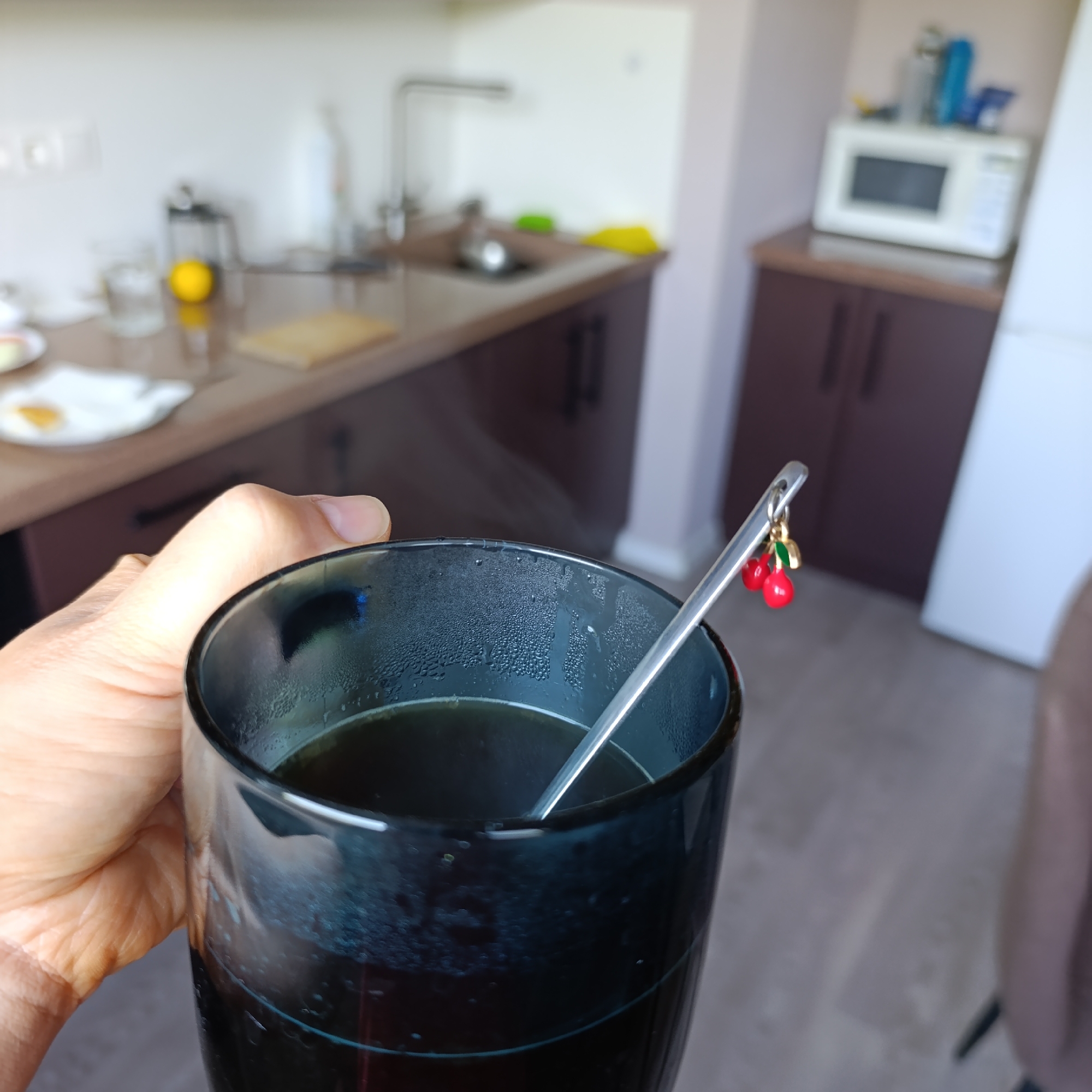 A person holds a dark, partially condensed glass filled with a dark liquid, possibly coffee or tea, with a silver spoon resting inside. The spoon is decorated with a charm of two red cherries. In the blurred background, a kitchen setting is visible. It includes a countertop with a sink, faucet, a cutting board, and some items including a lemon. To the right, there is a counter with dark wooden cabinets, topped with a white microwave and other items. The location is not discernible from the image and cannot be identified as Марковский, Russia.