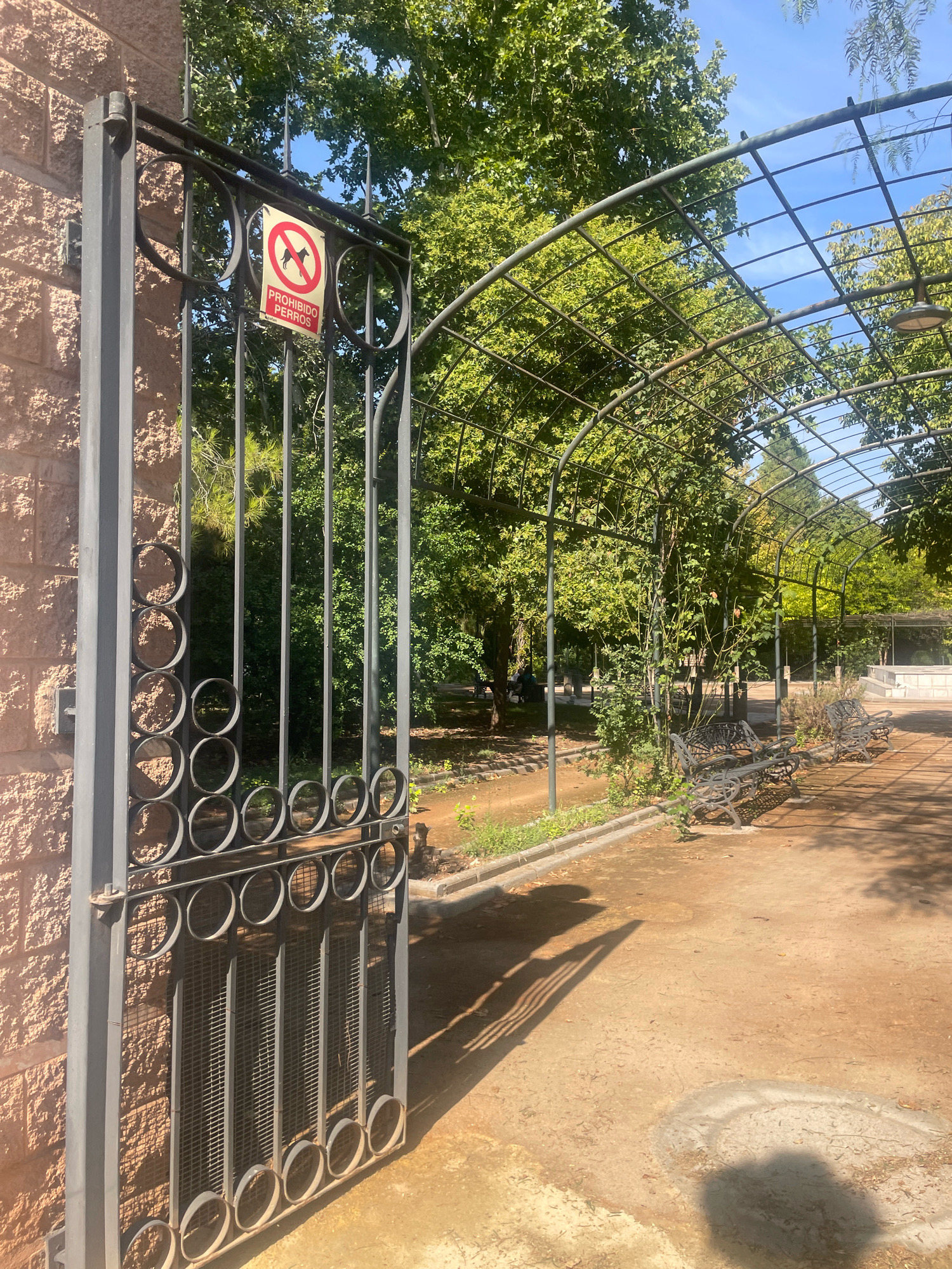 The image depicts an open wrought-iron gate leading into a park or garden area in Atarfe, Spain. The gate is on the left side of the frame and is attached to a textured brick wall. A sign on the gate displays a red circle with a line through it, indicating "Prohibido Perros," which translates to "No Dogs Allowed." Beyond the gate, a pathway of dirt and gravel leads through an arched structure covered in green foliage. Several empty, ornate benches are visible along the path. The park is filled with lush green trees and foliage, suggesting it is a warm, sunny day. The lighting indicates it is daytime, with shadows cast on the ground by the gate and the overhead structure. There are no people clearly visible in the image.