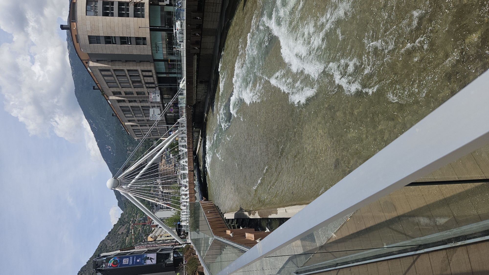 This image captures a vibrant urban scene in Andorra la Vella, Andorra, on a clear, sunny day with some scattered clouds. The primary subject is the Valira river, flowing from left to right, characterized by visible rapids and a clear, shallow appearance with a light brown, rocky bed.
In the foreground, a modern pedestrian walkway is visible, featuring a grey metal railing and a transparent glass barrier extending diagonally from the lower left to the mid-right. The walkway surface appears to be composed of light brown wooden planks.
Spanning the river in the midground is the distinctive Pont de la Rotonda (also known as Pont de París), a cable-stayed bridge. This bridge is architecturally notable for its central white spherical element from which numerous white cables radiate downwards to support the bridge deck. Large white capital letters on the bridge's framework spell out "ANDORRA LA VELLA." The riverbanks feature a mix of modern retaining walls, some appearing to be wooden or metallic on the left, and concrete or stone on the right.
In the background, on either side of the river and bridge, are multi-story urban buildings. To the left, a tall, dark building features a large vertical digital display with colorful graphics. Behind this, a steep, tree-covered mountain rises. To the right, a large, modern, curved building with numerous windows on multiple floors is prominent, with "MANGO OUTLET" signage visible on its ground level. A street runs in front of this building, with several white cars parked or in motion. Beyond the buildings, another large, forested mountain range dominates the horizon under a partially cloudy blue sky. A few indistinct figures are visible on the bridge and streets.
