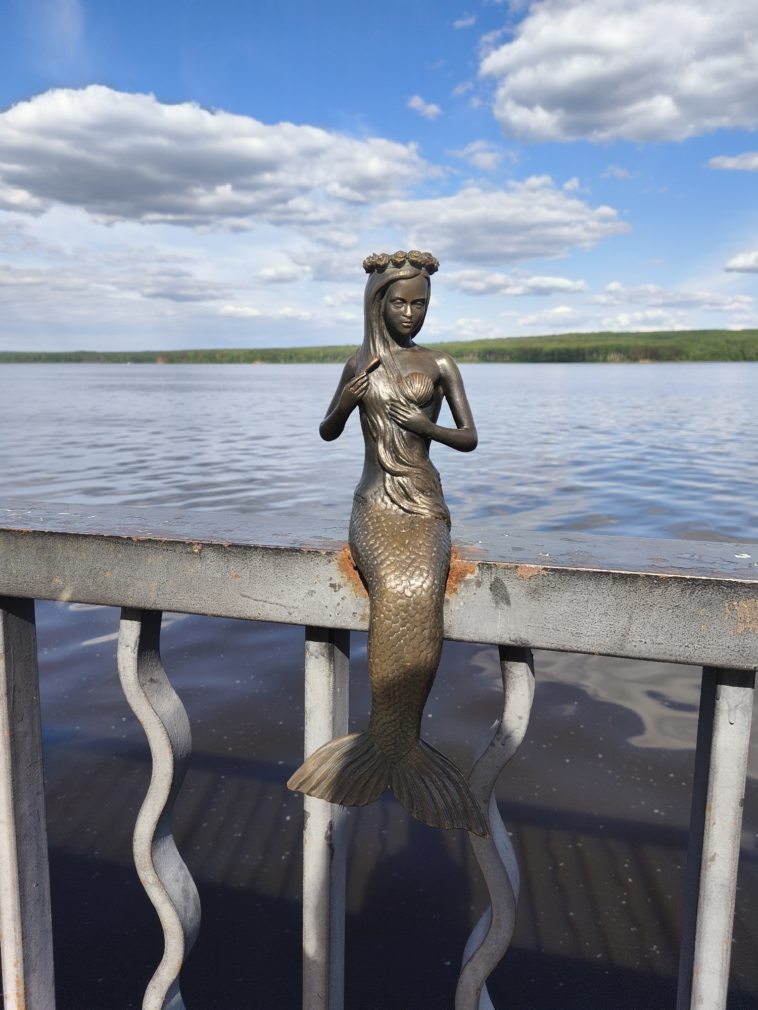 The image presents a bronze statue of a mermaid positioned on a metal railing, overlooking a body of water in the city of Chaikovsky, Russia. The mermaid statue, facing forward, features a crown of flowers on her head, long hair cascading down her shoulders, and her hands clasped over a seashell on her chest. Her scaled tail extends downwards, resting on the top bar of the railing.
The railing, appearing weathered and metallic gray with areas of rust and textured surface, spans horizontally across the lower mid-ground of the frame. It consists of a thick top bar supported by vertical posts and undulating, wave-like decorative elements. Below the railing, the dark, reflective surface of the water is visible, showing faint ripples.
In the mid-ground, beyond the railing, is a wide expanse of water, displaying a choppy surface with varying shades of blue and gray, indicating movement. A distant treeline, composed of dense green foliage, forms the horizon line across the far bank of the water body. The sky above is largely clear blue with several scattered, puffy white cumulus clouds. The scene is brightly illuminated by natural daylight, casting subtle shadows.