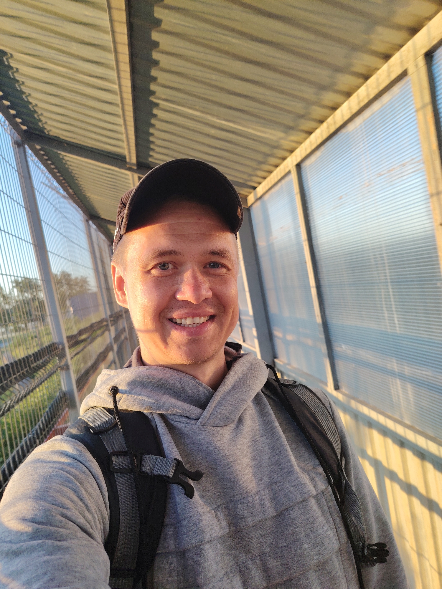 A man in a grey hoodie and a dark baseball cap takes a selfie. He is standing on a pedestrian overpass with a corrugated metal roof and clear, corrugated plastic walls. A metal fence runs along the left side of the overpass, separating it from a grassy area with trees. The sun is low in the sky, casting long shadows and highlighting the texture of the overpass's roof and walls. The overpass appears to be located in a semi-urban or suburban area, possibly near a road or railway line. The overall lighting suggests it is either early morning or late afternoon.