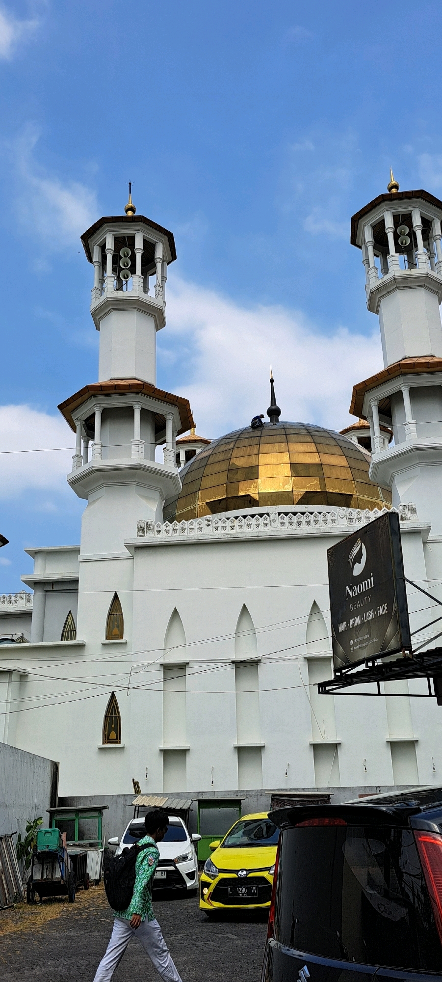 The image captures the exterior of a mosque with a prominent golden dome and two white minarets against a bright blue sky with scattered clouds. The mosque's white facade features arched windows. In the foreground, a person wearing a green patterned shirt and grey pants walks from left to right, carrying a black backpack. To the right of the mosque, a black billboard for "Naomi Beauty" is visible, advertising hair, brow, lash, and face services. Several cars are parked in front of the mosque, including a white car and a yellow car with a license plate reading "L 1290 VV". The scene appears to be in a city location, possibly Petungasri, Indonesia, given the context. Electrical wires run across the lower portion of the image.