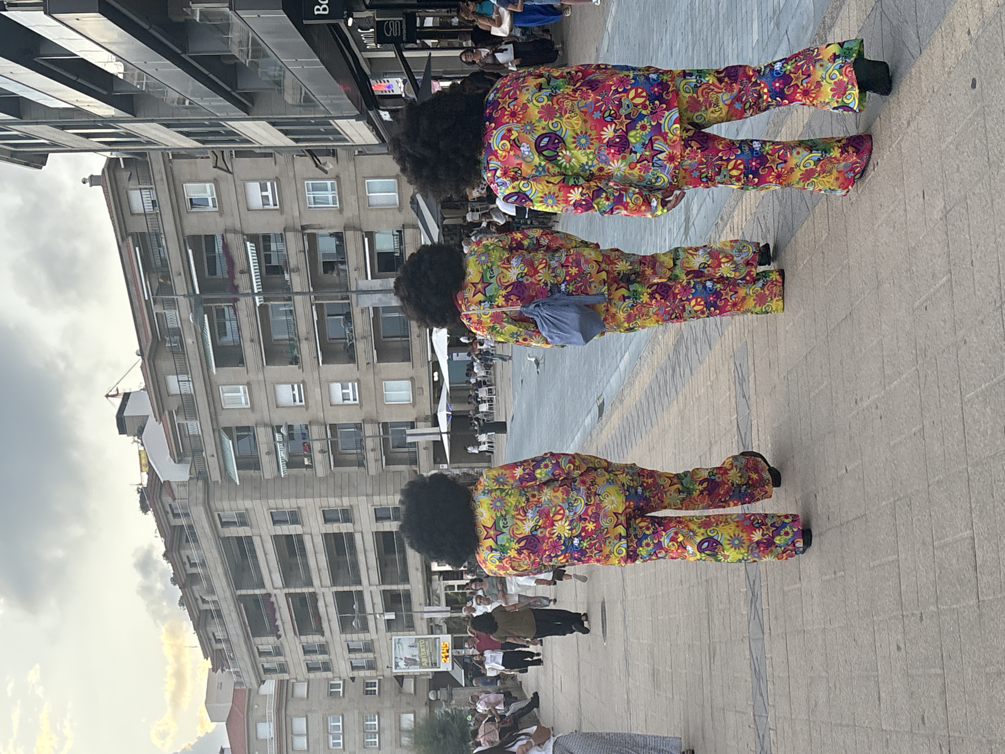 Three individuals wearing matching colorful, psychedelic-patterned outfits, complete with large afro wigs, are walking away from the camera on a paved walkway in Sanxenxo, Spain. The outfits feature a vibrant mix of patterns including peace signs, flowers, and stars in yellow, pink, green, blue, and orange. The person in the center is carrying a blue drawstring bag. They are walking on a sidewalk that is bordered by buildings. To the left, a multi-story building with balconies and apartments can be seen. To the right, a commercial building with storefronts and an awning is visible. In the background, other people are walking and sitting at outdoor tables under umbrellas, suggesting a public area or promenade. The sky is cloudy with patches of sunlight.
