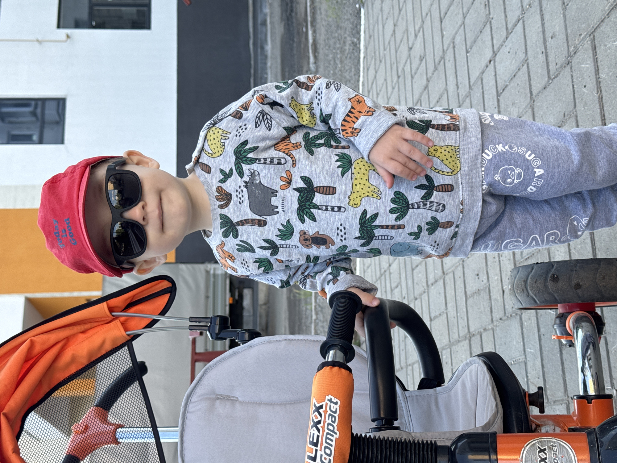 A young child stands outdoors next to an orange and gray tricycle-stroller in Izhevsk, Russia. The child, appearing to be a toddler, is dressed in a gray long-sleeved top adorned with a colorful jungle animal pattern, including leopards, tigers, rhinos, and palm trees. They wear matching gray sweatpants, dark sunglasses, and a red baseball cap with some faint white text, possibly "GOOD DOG." The child's right hand rests on the black handlebar of the tricycle, which features an orange handle cover with "LEXX compact" visible in white text. The gray sweatpants also display white text and a duck logo that reads "DUCK SUGAR DUCK." The child looks towards the camera with a neutral expression. The background shows a light-colored building with dark windows and a paved ground made of rectangular gray blocks, indicating an urban or suburban outdoor setting. The scene is brightly lit, suggesting daytime.