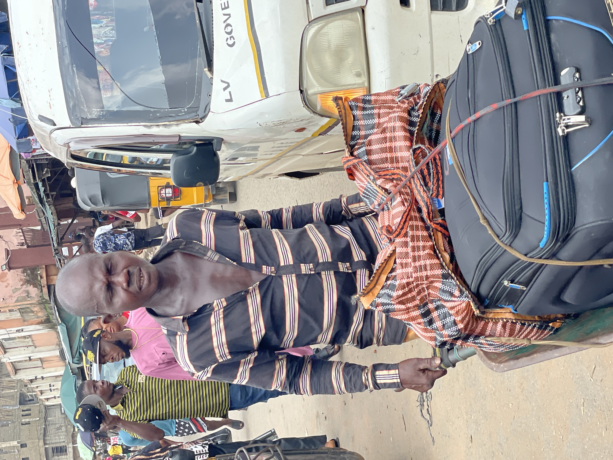 The image presents a busy street scene in the city of Unknown, Unknown. In the foreground, a dark-complexioned man with a bald head is positioned slightly left of center, appearing to push a two-wheeled metal handcart. He wears a long-sleeved collared shirt with alternating black and light-colored horizontal stripes, partially unbuttoned to reveal his chest. His eyes are partially closed, and his mouth is slightly open, revealing some teeth. His left hand firmly grips the handle of the handcart.
The handcart carries two items secured with thin ropes: a black, wheeled travel suitcase or large bag positioned at the bottom, and on top, a larger, soft bag made of a woven plastic material featuring a red, black, and white plaid pattern.
In the midground, to the left and behind the primary subject, several other men are visible. One man wears a yellow and black striped polo shirt and holds a black and yellow baseball cap in his right hand. Further back, another man with a beard is seen wearing a pink long-sleeved shirt and a black baseball cap worn backward, along with a gold chain. Other indistinct figures are visible, suggesting a crowd.
On the right side of the image, the front portion of a white van or minibus is visible, facing left. The vehicle shows signs of wear and has a crack across its windshield. The letters "LV GOVER" are printed in black on its front panel. Behind the white van, part of a yellow three-wheeled auto-rickshaw is discernible.
The background consists of multi-story buildings in varying conditions lining the street, some with visible tarpaulin awnings or umbrellas overhead. The street surface appears to be a mix of dirt and worn pavement.