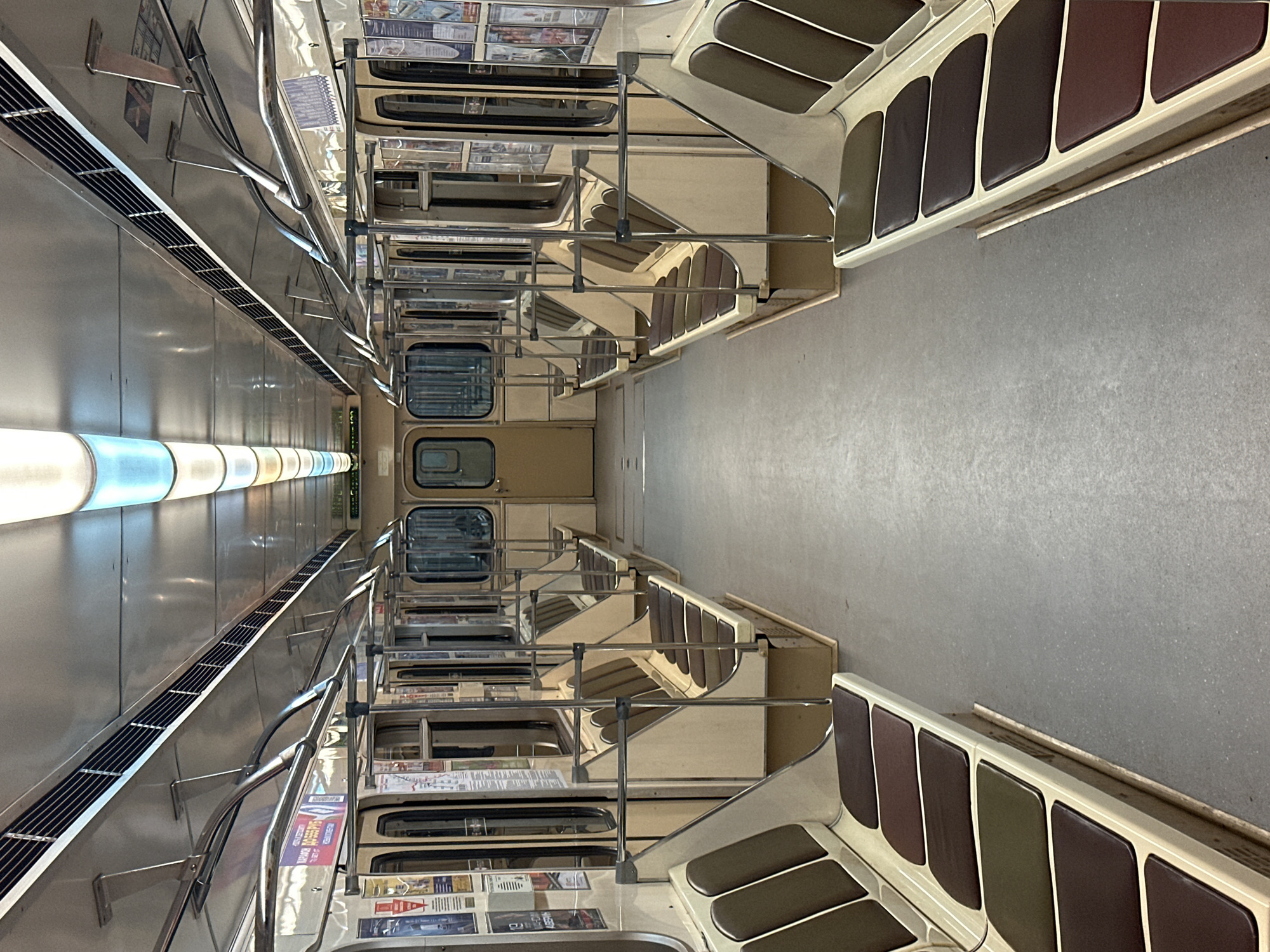 The image shows the interior of an empty subway car, likely in Minsk, Belarus, given the context of the prompt. The car features rows of beige and brown upholstered seats along both sides, facing inwards. Above the seats, silver poles run parallel to the length of the car, offering passengers something to hold onto. The ceiling is a reflective silver material, illuminated by long, rectangular light fixtures that emit a cool, white light. Various posters and advertisements are visible on the walls above the windows. The floor is a plain, grey, textured surface. The overall impression is one of a clean, well-maintained, but dated public transportation vehicle.