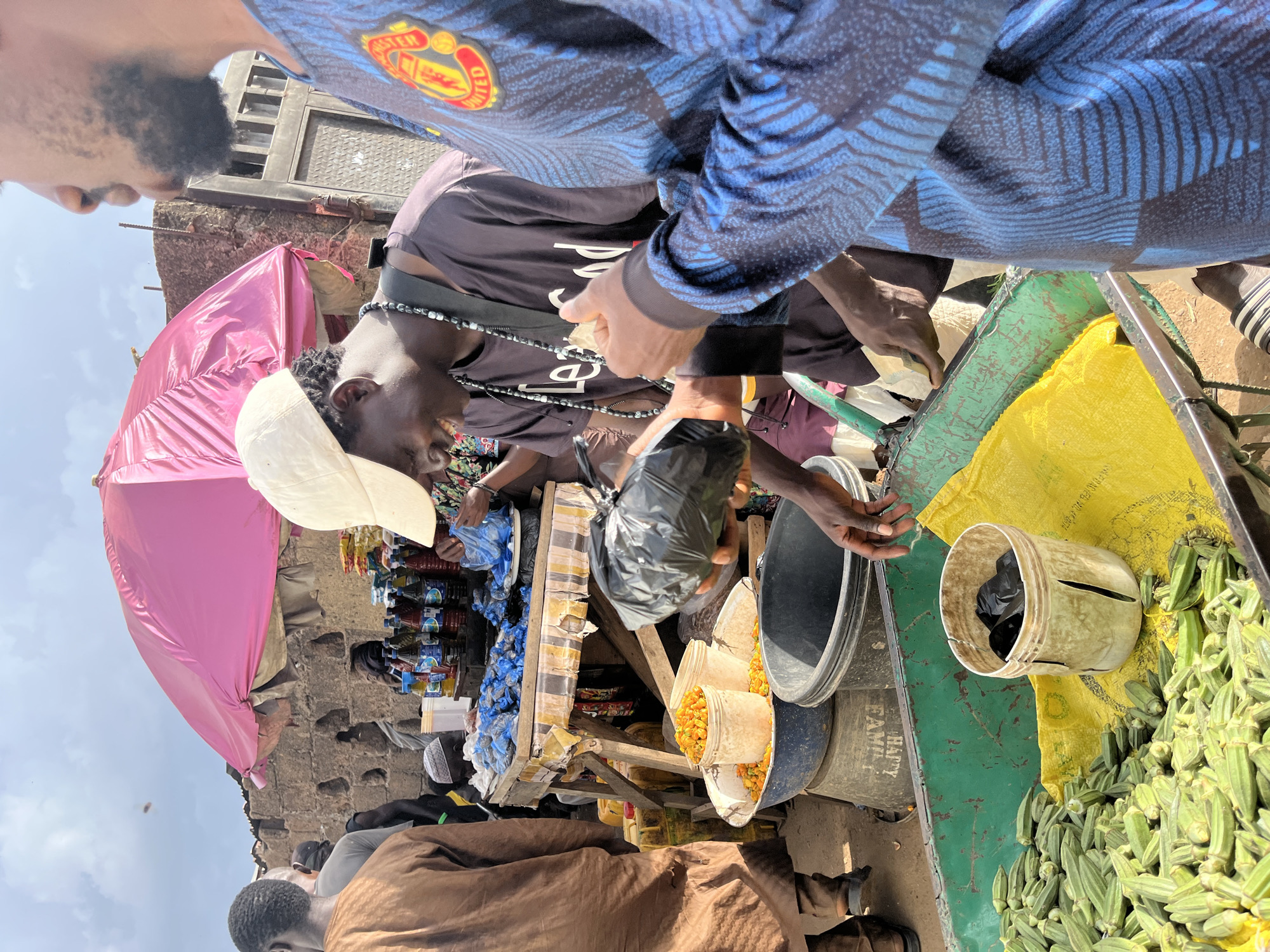 The image depicts a bustling outdoor market scene in Rafin Par, Nigeria. In the foreground, a vendor wearing a dark t-shirt and a white cap is handing a plastic bag to a customer, who is partially visible on the right, wearing a blue shirt with a Manchester United logo. The vendor is smiling and appears to be completing a transaction. Behind them, a large pink umbrella provides shade for a stall laden with bottled drinks and other goods. In the middle ground, a wheelbarrow overflowing with fresh okra is visible, suggesting produce is a primary commodity. Several people are milling about in the background, engaged in market activities. The overall environment suggests a typical lively Nigerian marketplace.