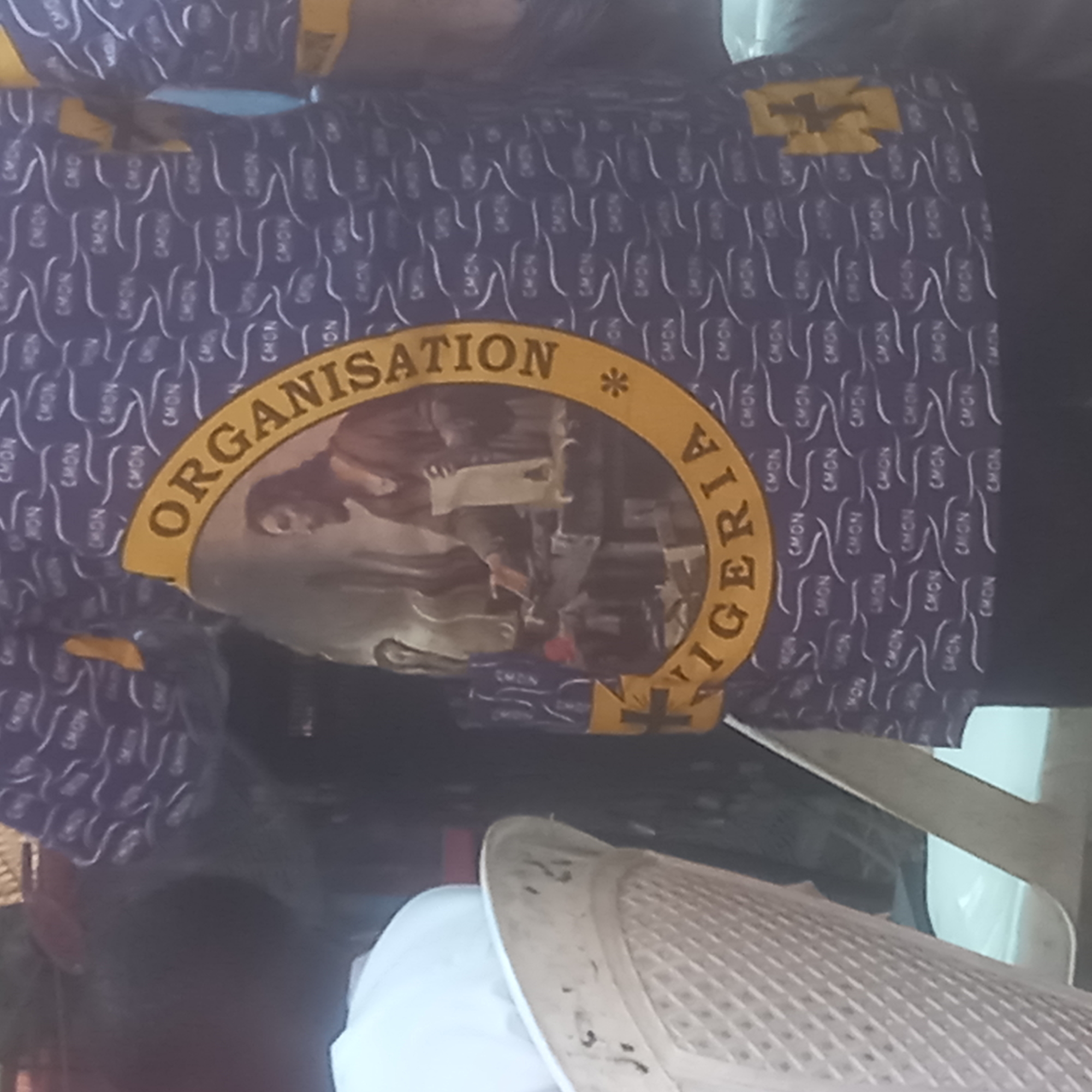 The image shows a piece of dark blue fabric with repeating patterns of the letters "CMON". On the fabric is a circular design in gold, containing an image of a group of people. Above and below the image, the gold circle contains the words "ORGANISATION" and "NIGERIA," separated by a star. The background is blurred. The location is Barden Yana, Nigeria.