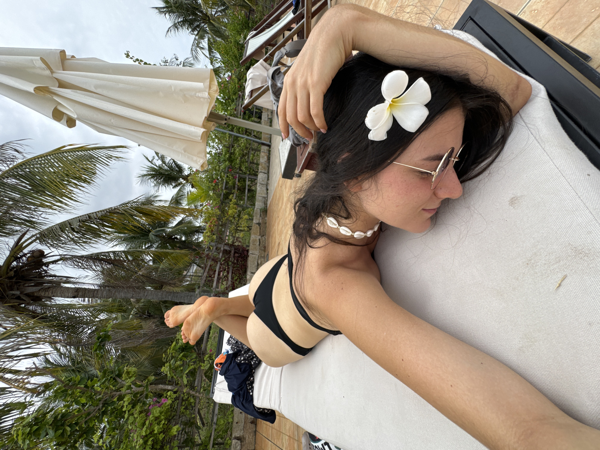 A young woman is captured in a relaxed pose, lying on her stomach on a white lounge chair in an outdoor setting. She has long dark hair styled with a white plumeria flower tucked behind her left ear. She wears small, round, rose-tinted sunglasses and a seashell necklace. Her eyes are closed, suggesting she is resting or sunbathing. She is dressed in a black two-piece bikini. Her left arm is bent, with her hand resting near her head, while her right arm extends along the lounge chair.
The background reveals a tropical environment, consistent with Phu Thuy Ward, Vietnam, featuring numerous tall palm trees and abundant green foliage. A large, closed, off-white beach umbrella stands to her left. The sky is overcast, indicating a cloudy day. The ground around the lounge area appears to be paved with light-colored tiles. The scene conveys a sense of peaceful leisure and vacation. No visible text is legible in the image.